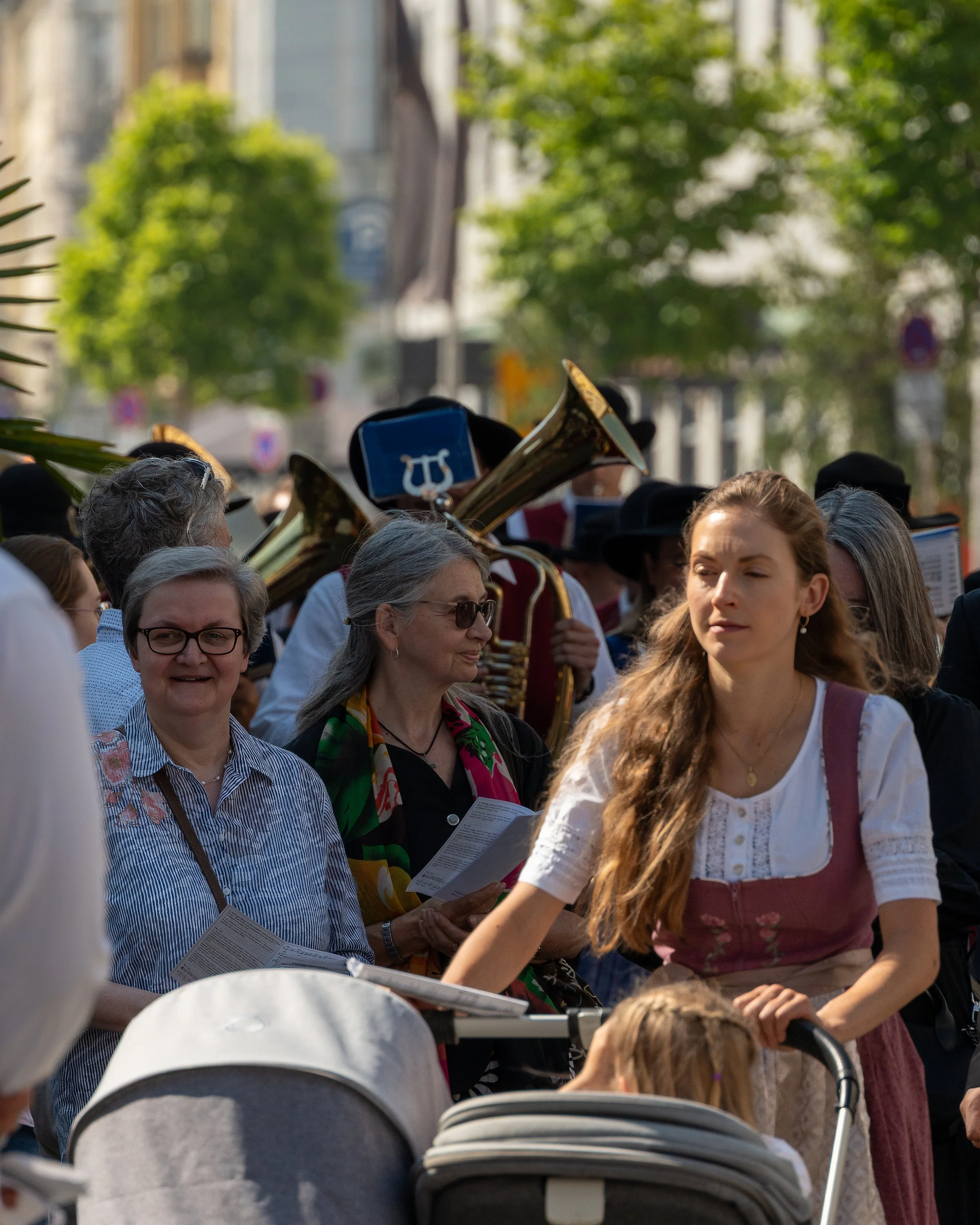 A crowded outdoor scene with people, some holding sheet music, and a brass band playing instruments in the background in a sunny, tree-lined city street.