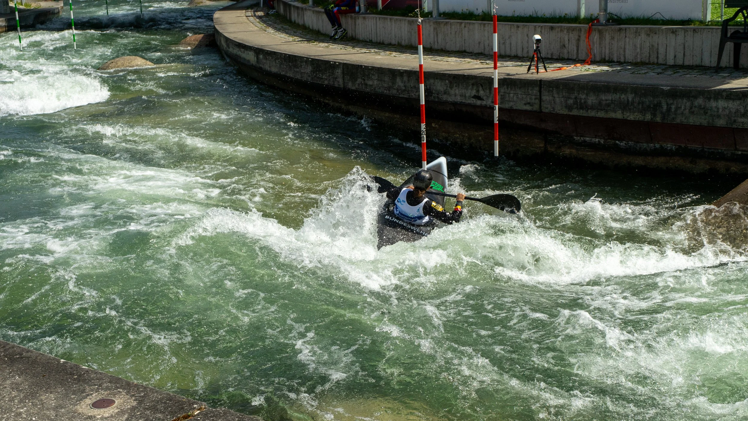 A kayaker in black and white gear navigating white water rapids in a man-made whitewater course with red and white gate poles, and a concrete wall and walkway along the side.