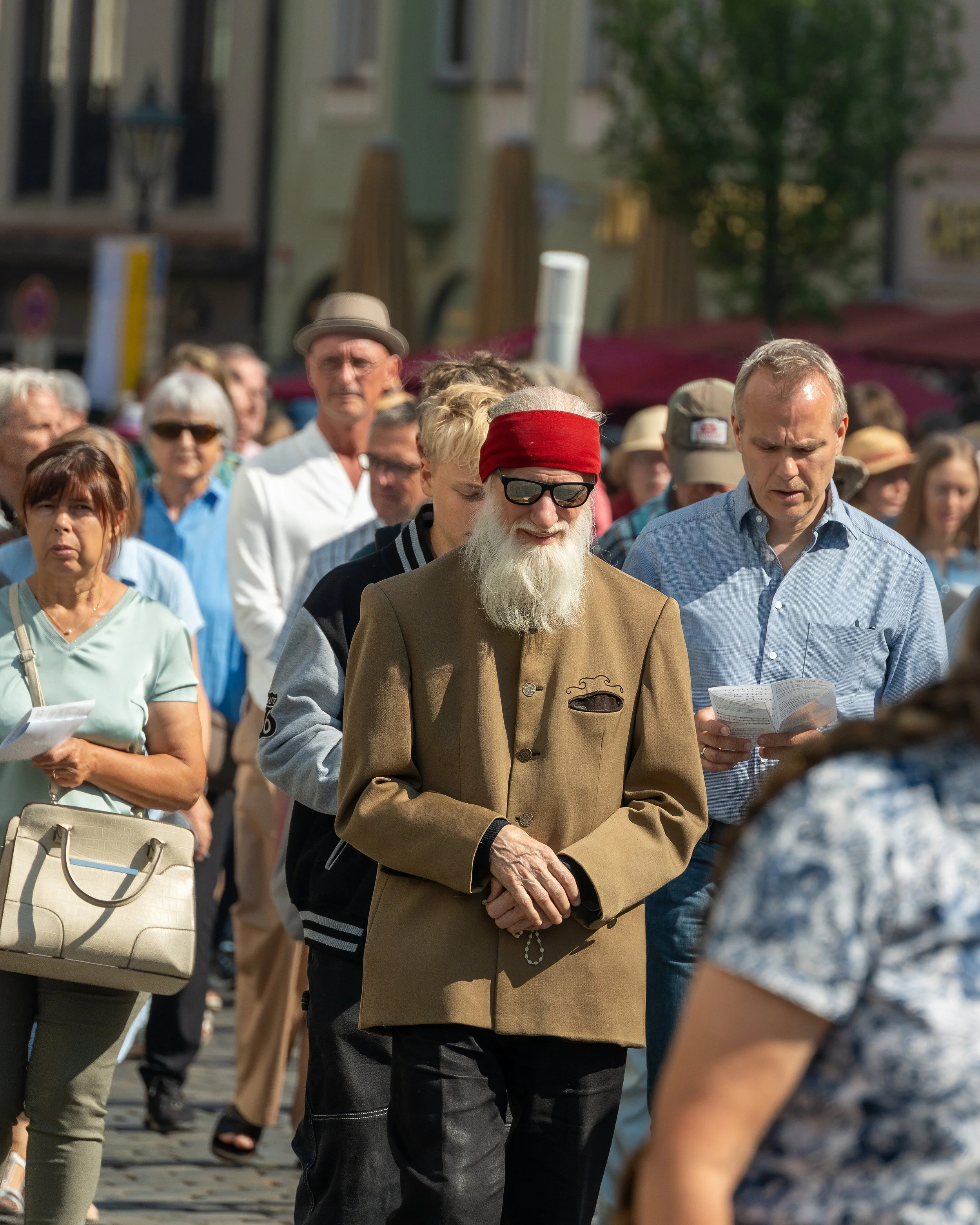 A crowd of people walking outdoors on a sunny day, with a man with a long gray beard, sunglasses, red headband, and tan jacket in the center.