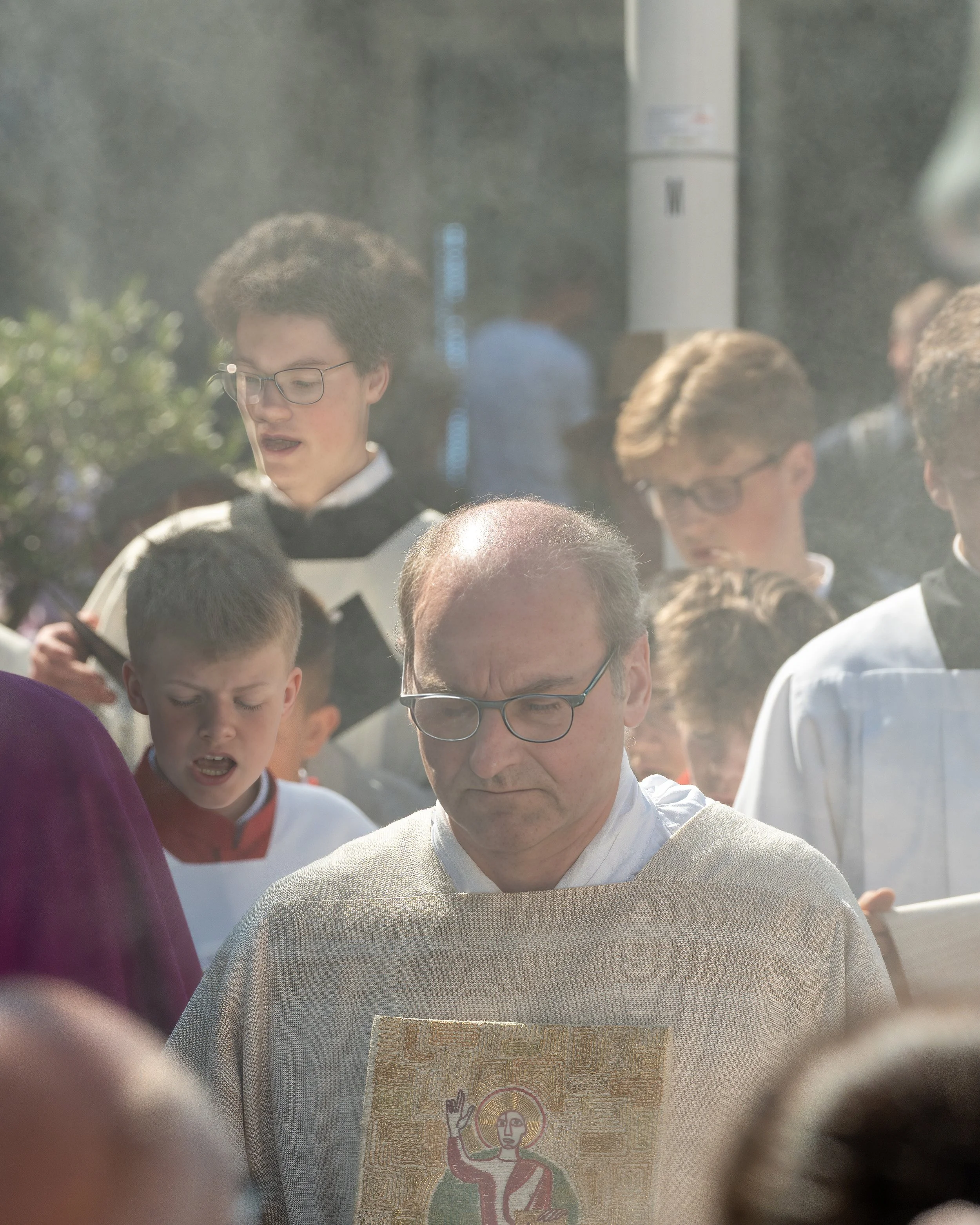 A group of people participating in a religious outdoor ceremony, with a priest holding an icon of a saint, and others with bowed heads and solemn expressions.