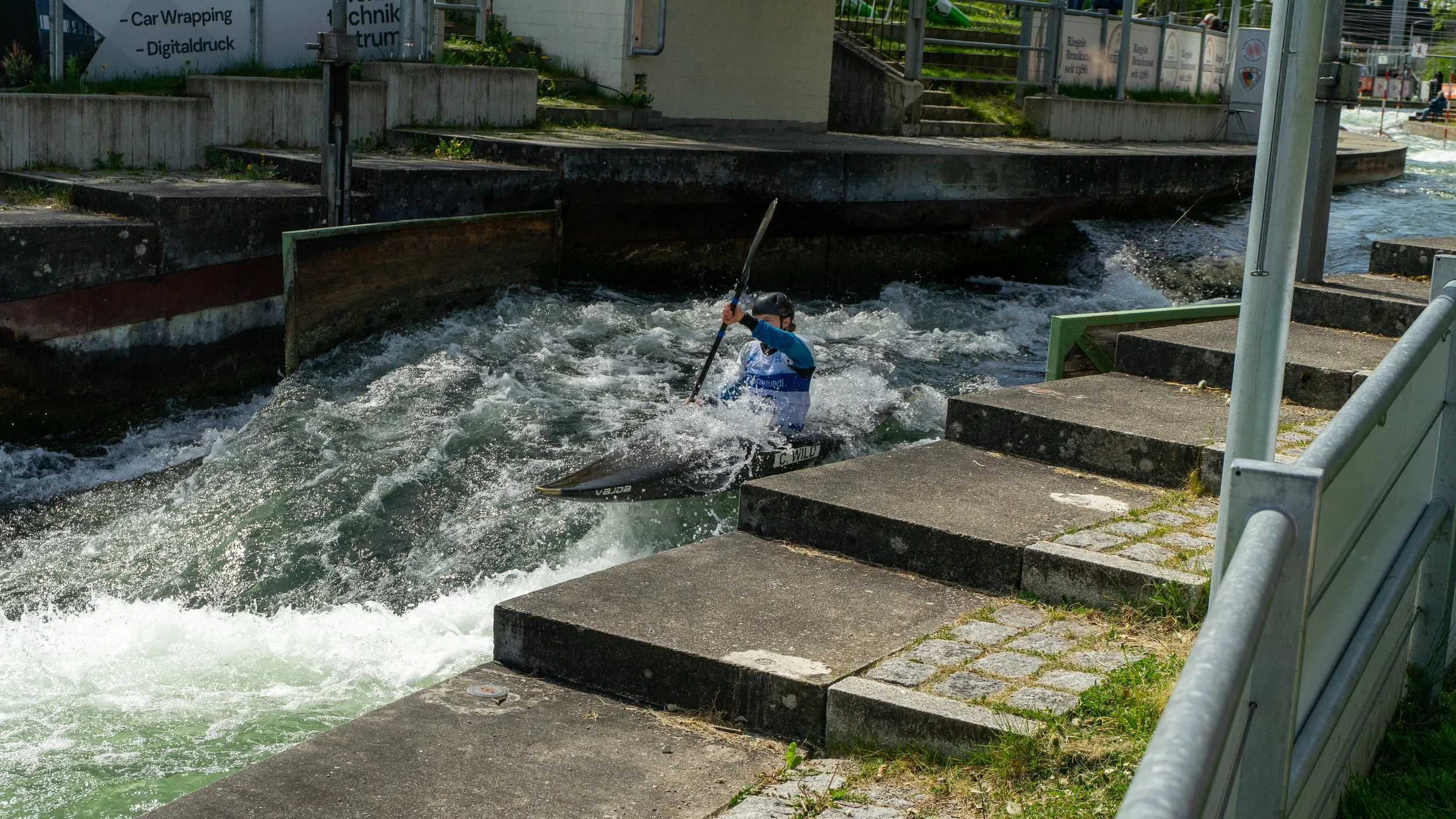A person kayaking in a narrow water channel with a concrete and metal railing on either side, near a set of stairs and a sign with German text.