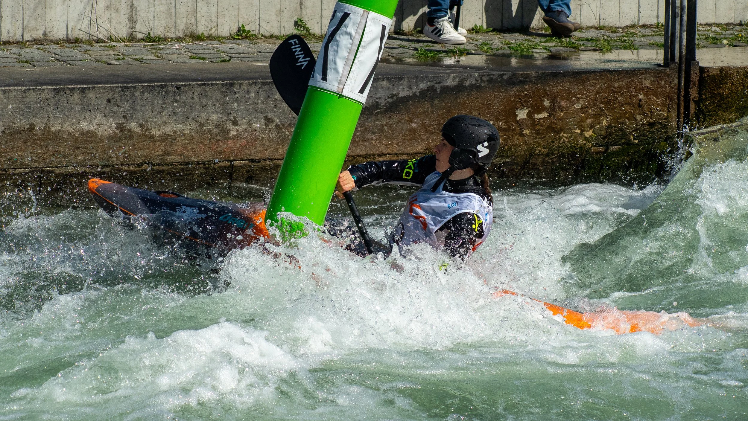 A person in a helmet and life jacket paddling a kayak in turbulent water near a concrete edge, with a green pole nearby.