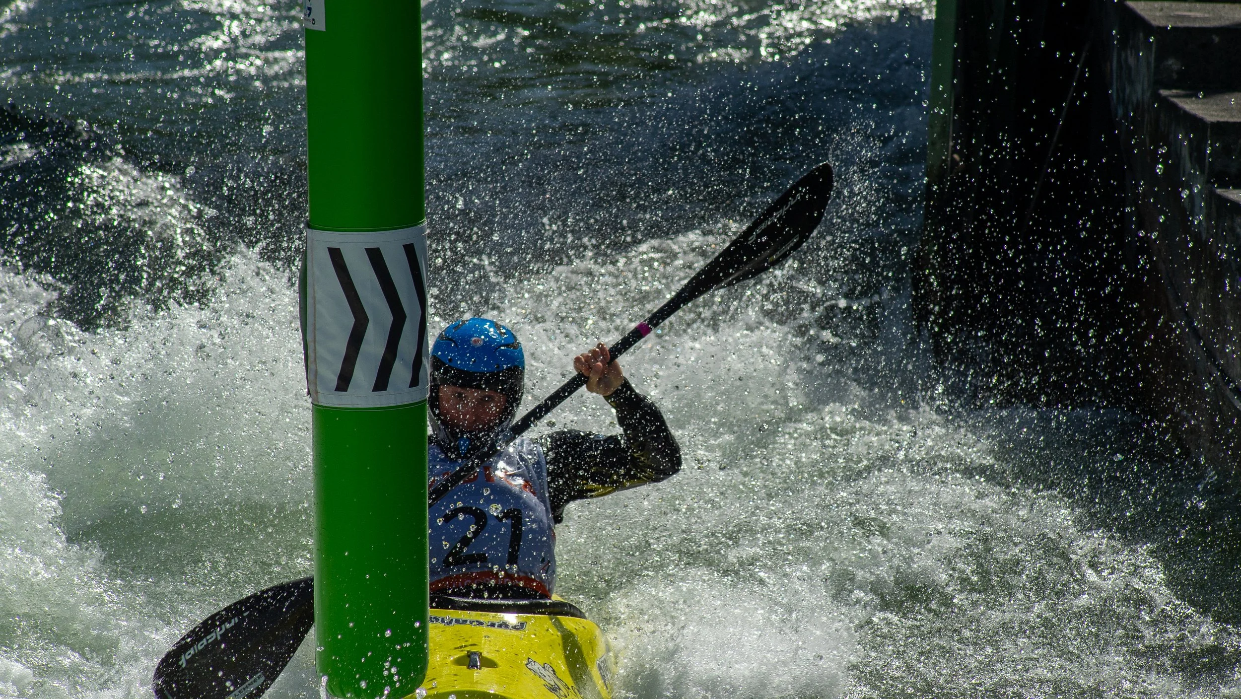 A person wearing a blue helmet and life jacket paddling a yellow kayak through whitewater rapids, with a large green marker pole in the foreground.