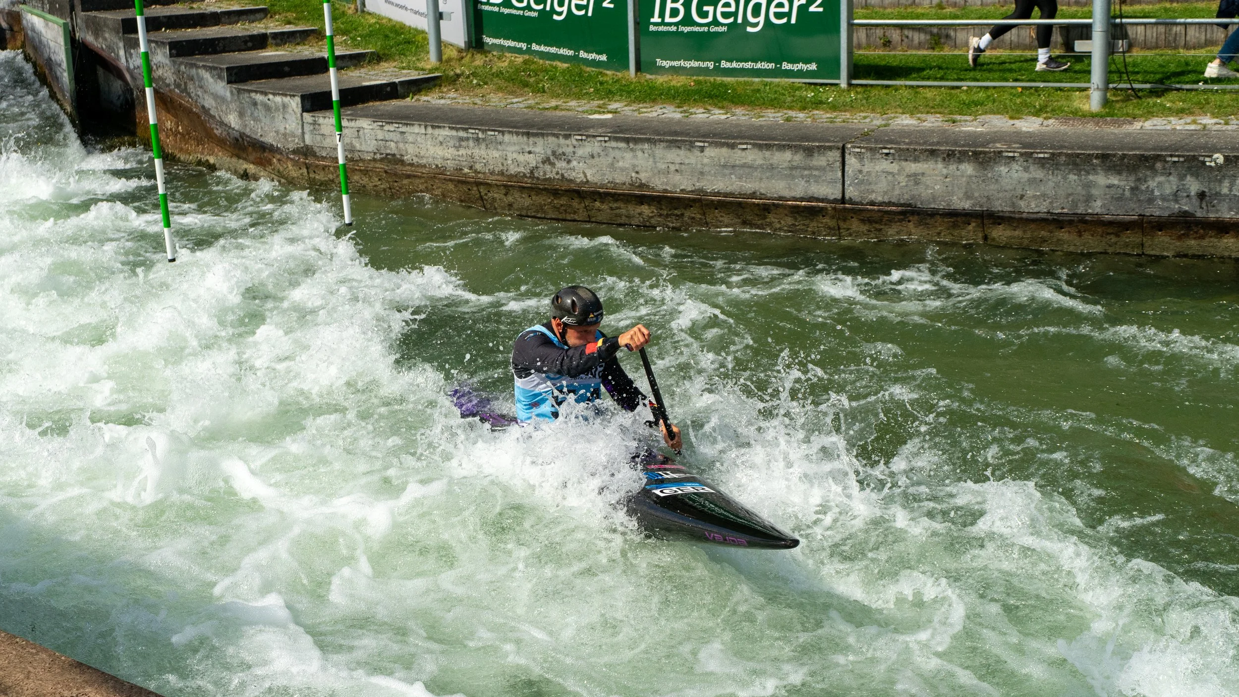 A person wearing a helmet and a life vest is kayaking through a whitewater course with turbulent water, approaching a hanging obstacle in a sports facility.