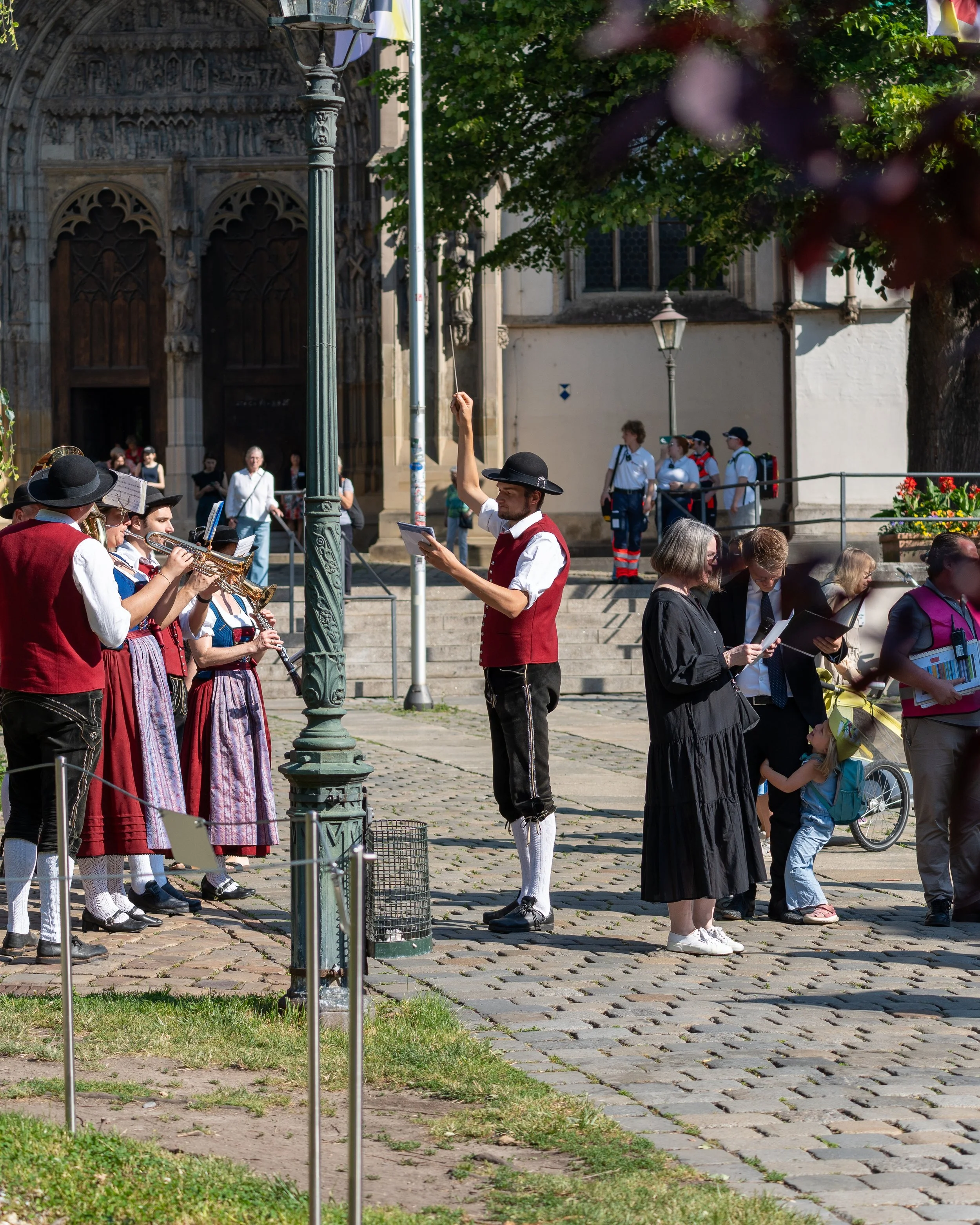 A group of people, including musicians dressed in traditional European attire, are gathered outdoors near a historic building. Some people appear to be engaged in a musical performance or rehearsal, while others are reading sheet music or observing.