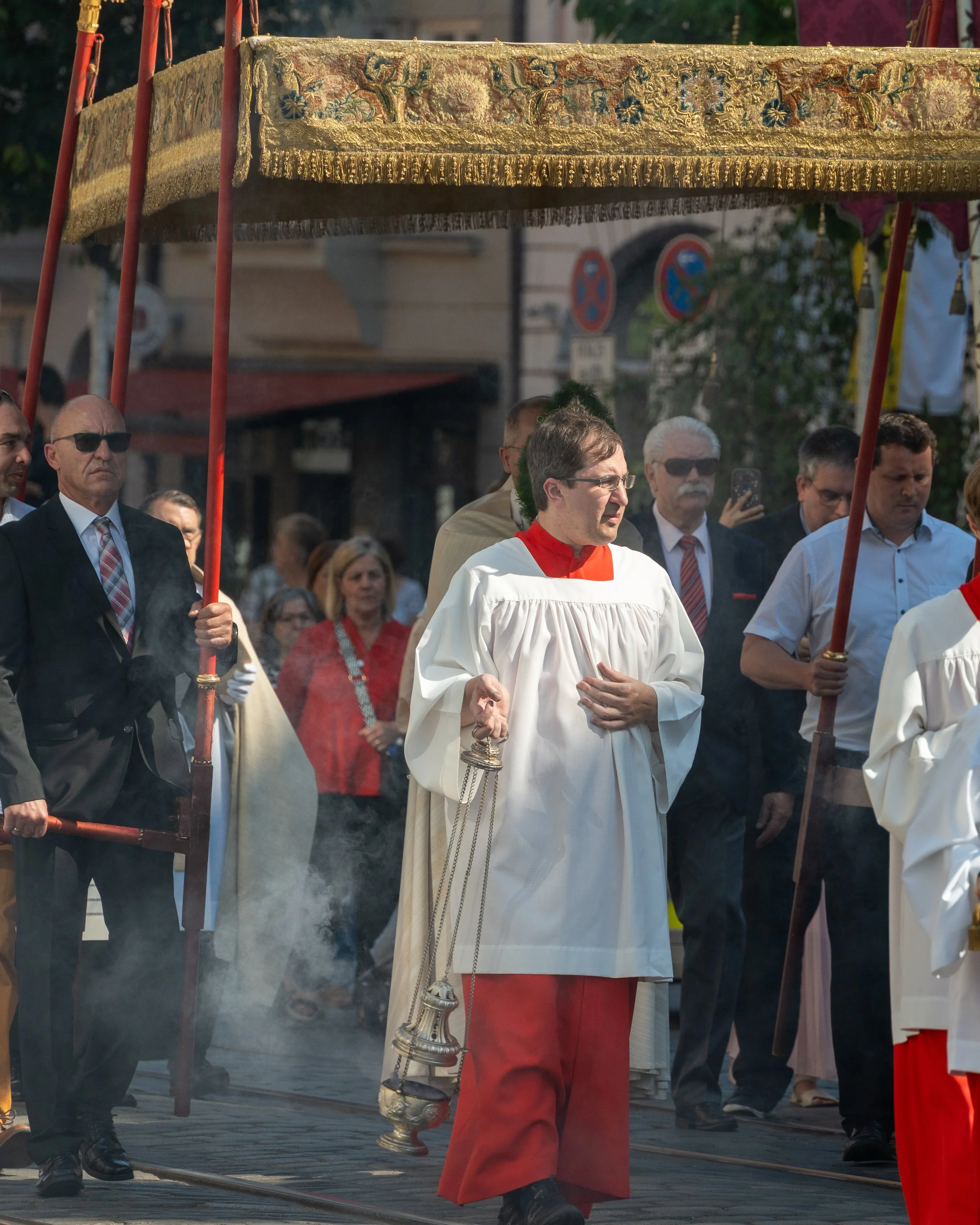 A religious procession with clergy and participants walking on a street in daytime, some holding a decorative canopy with ornate fabric, others carrying incense and dressed in ceremonial robes.