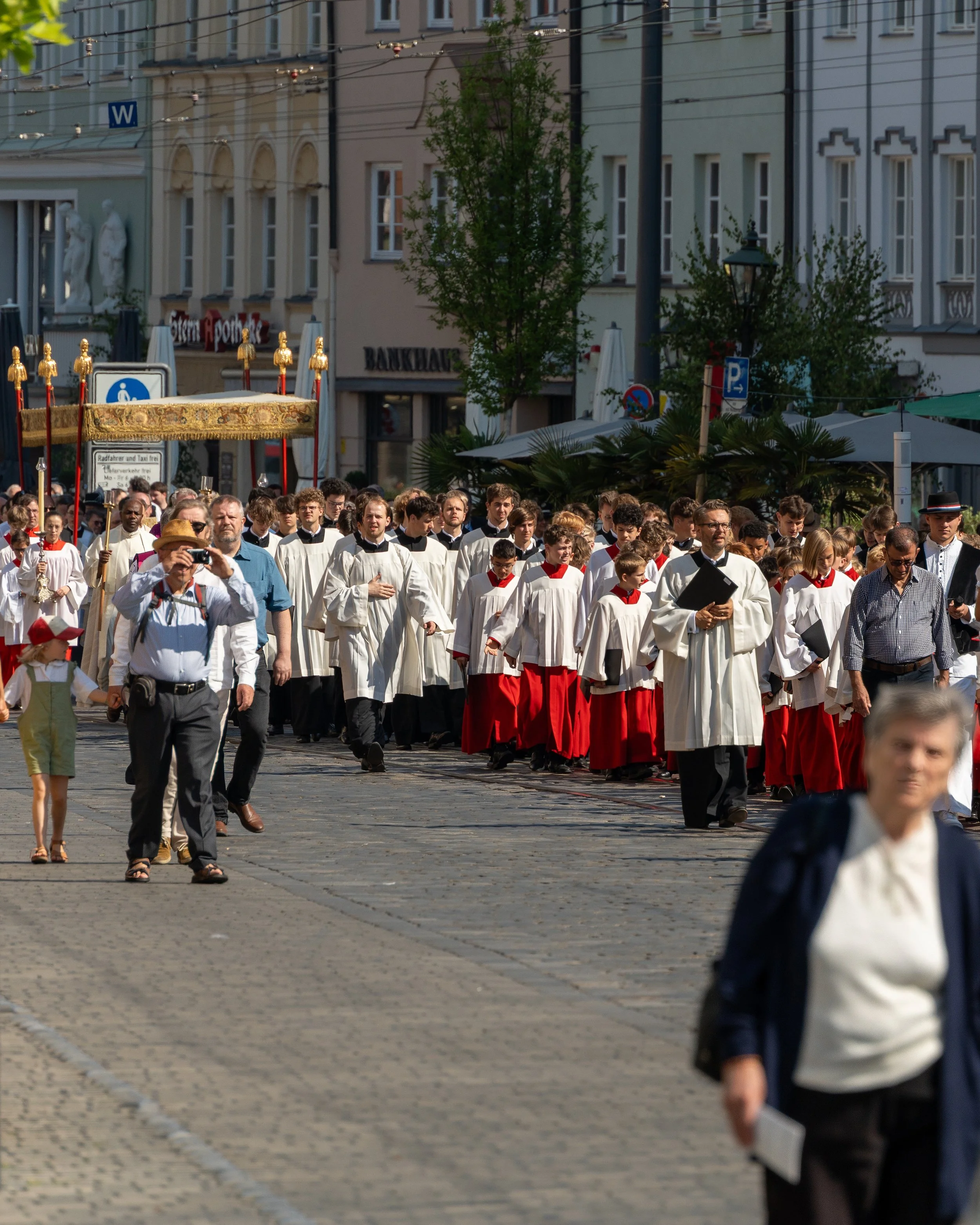 A religious procession with people dressed in white and red robes, walking on a cobblestone street in a European city, with some officials and spectators taking photos.