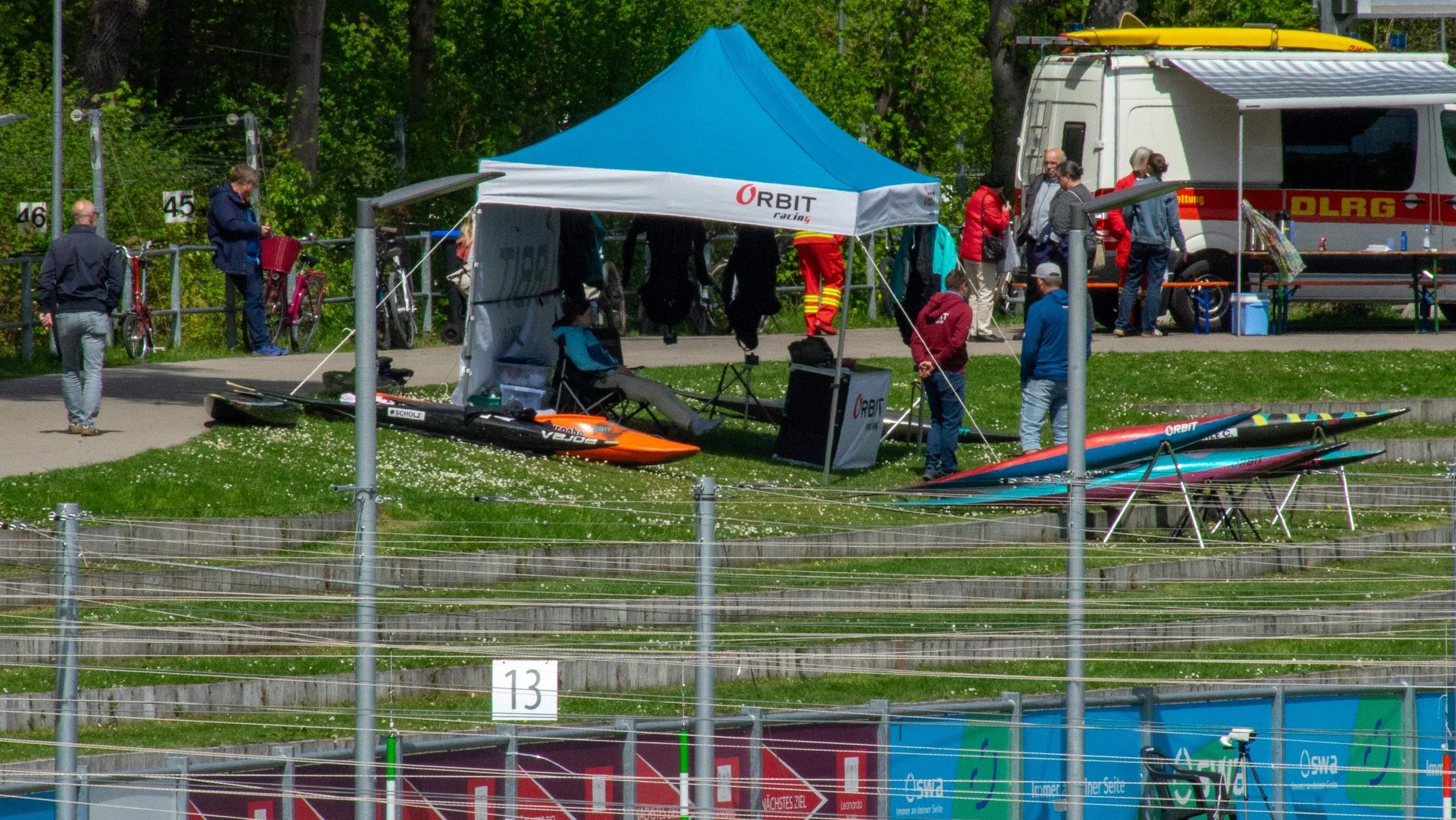 People gathered at a sports event with kayaks and paddleboards on the grass and tents set up, including a tent with Orbit Racing branding, near an emergency response vehicle.