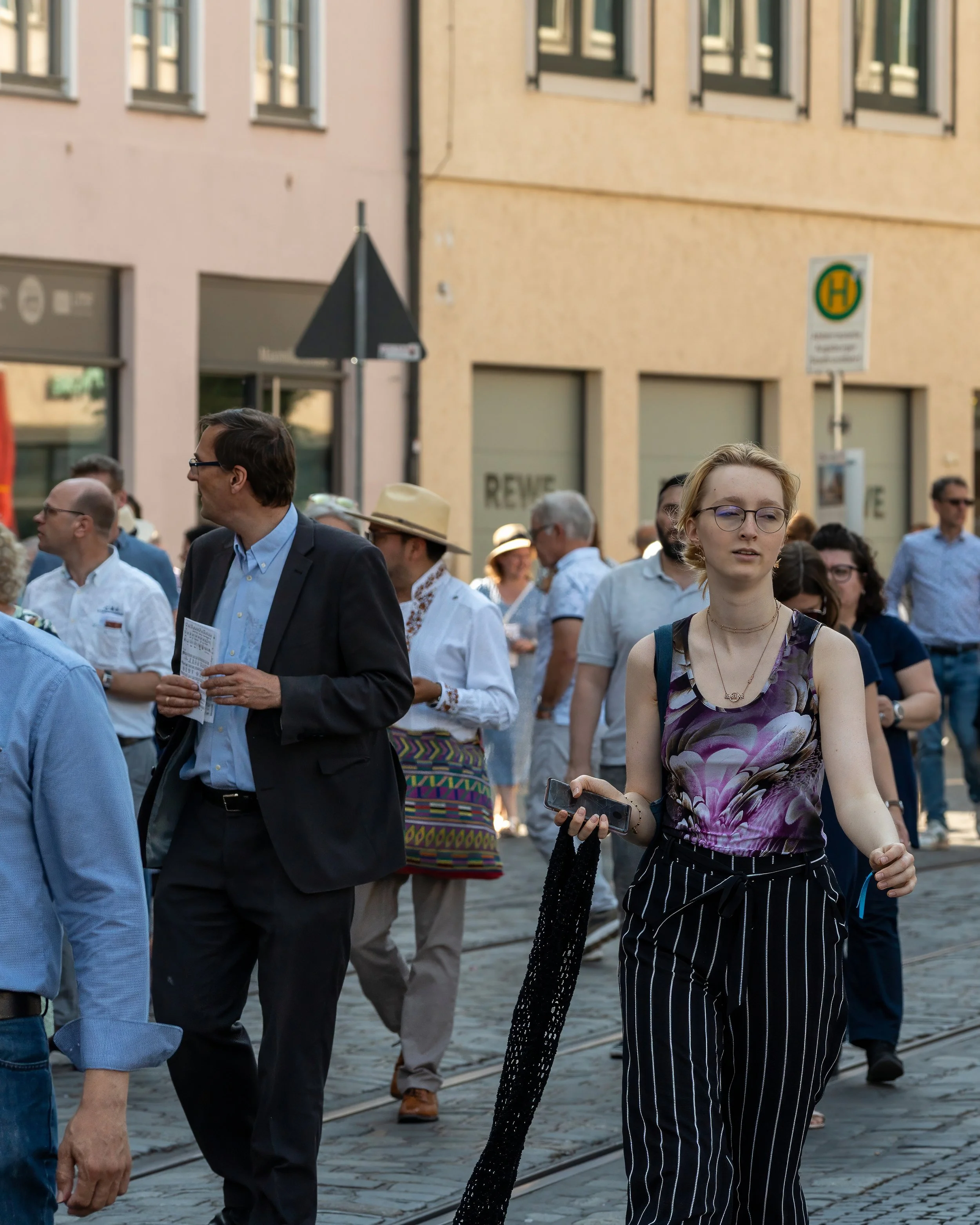 Crowd of people walking on a city street with buildings and a bus stop sign in the background.