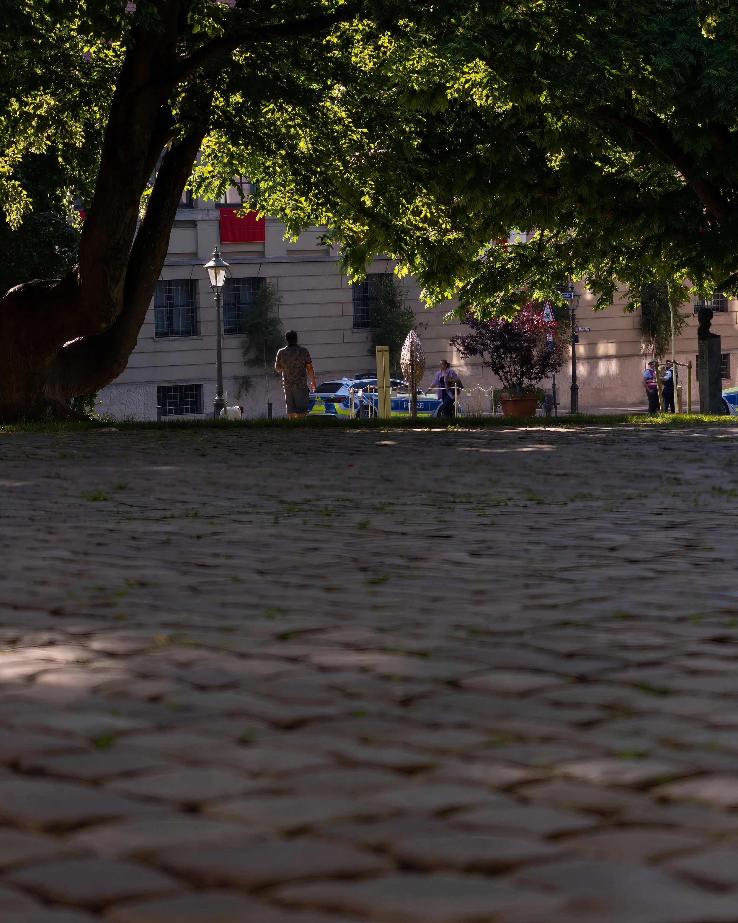 A low-angle view of a city park with cobblestone pavement, large tree with green leaves providing shade, and people walking along the sidewalk. A police car is parked in front of a beige building with windows, and a few individuals are seen near the