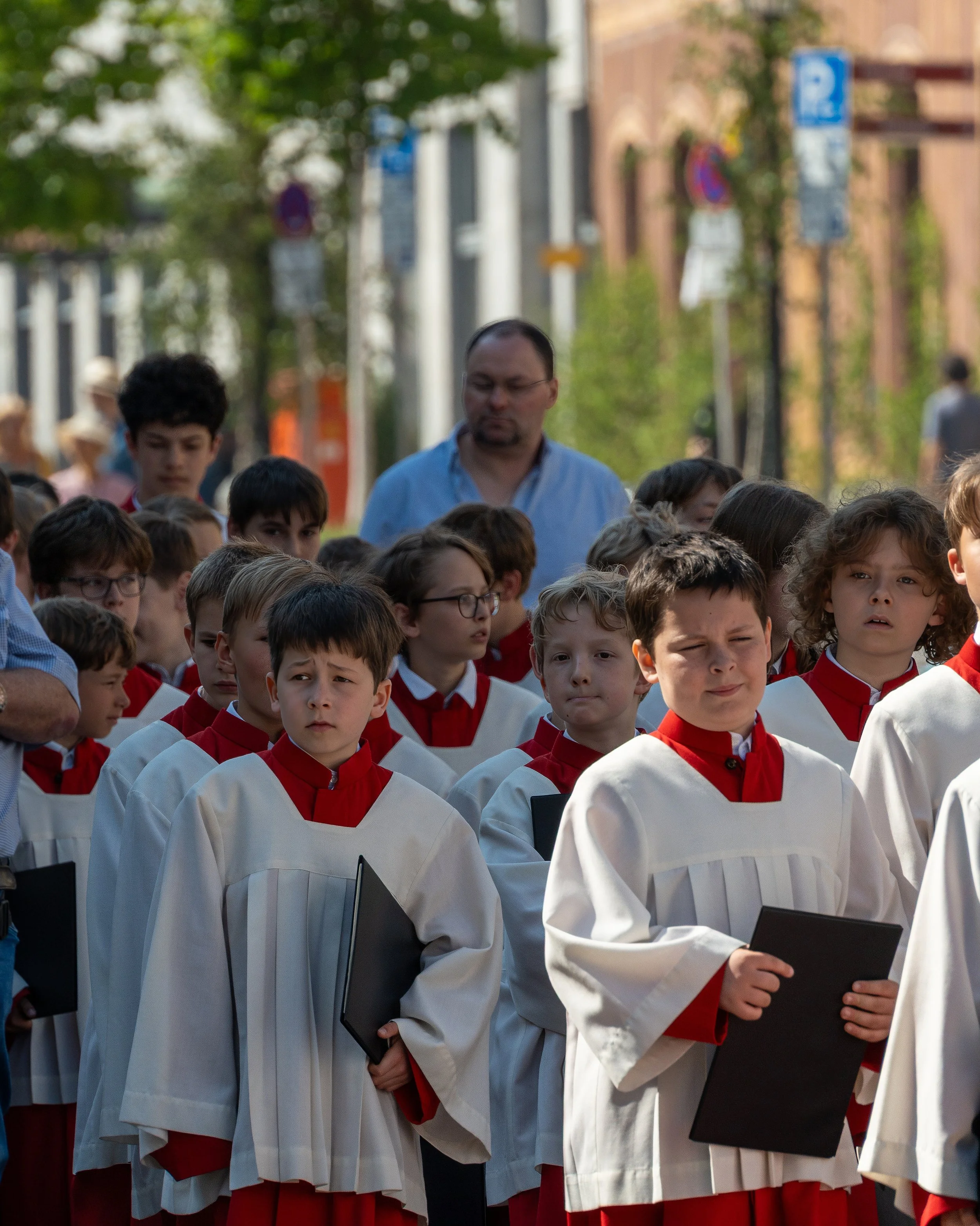 Children wearing white and red choir robes holding black folders, standing outdoors during daytime, with a man and other people in the background.