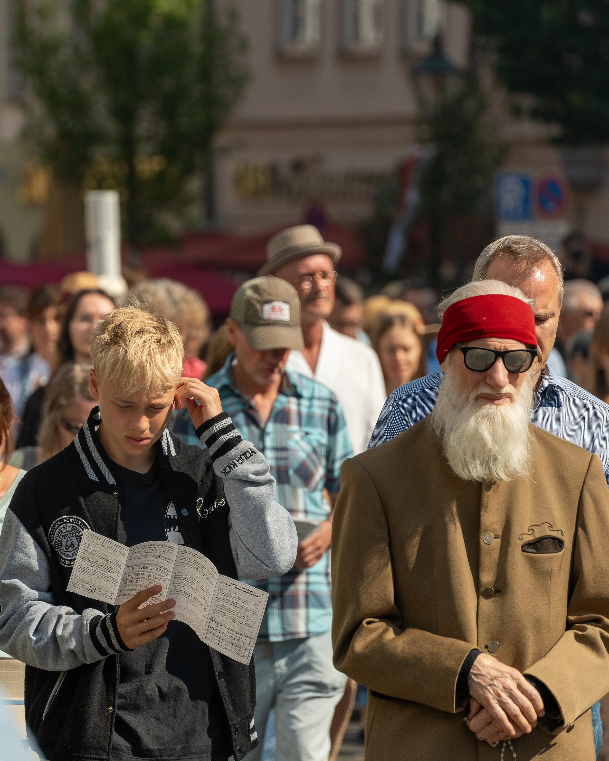 A crowd of people standing outdoors, several are looking down at papers or prayer books, with some wearing sunglasses and hats, in a sunny urban setting.