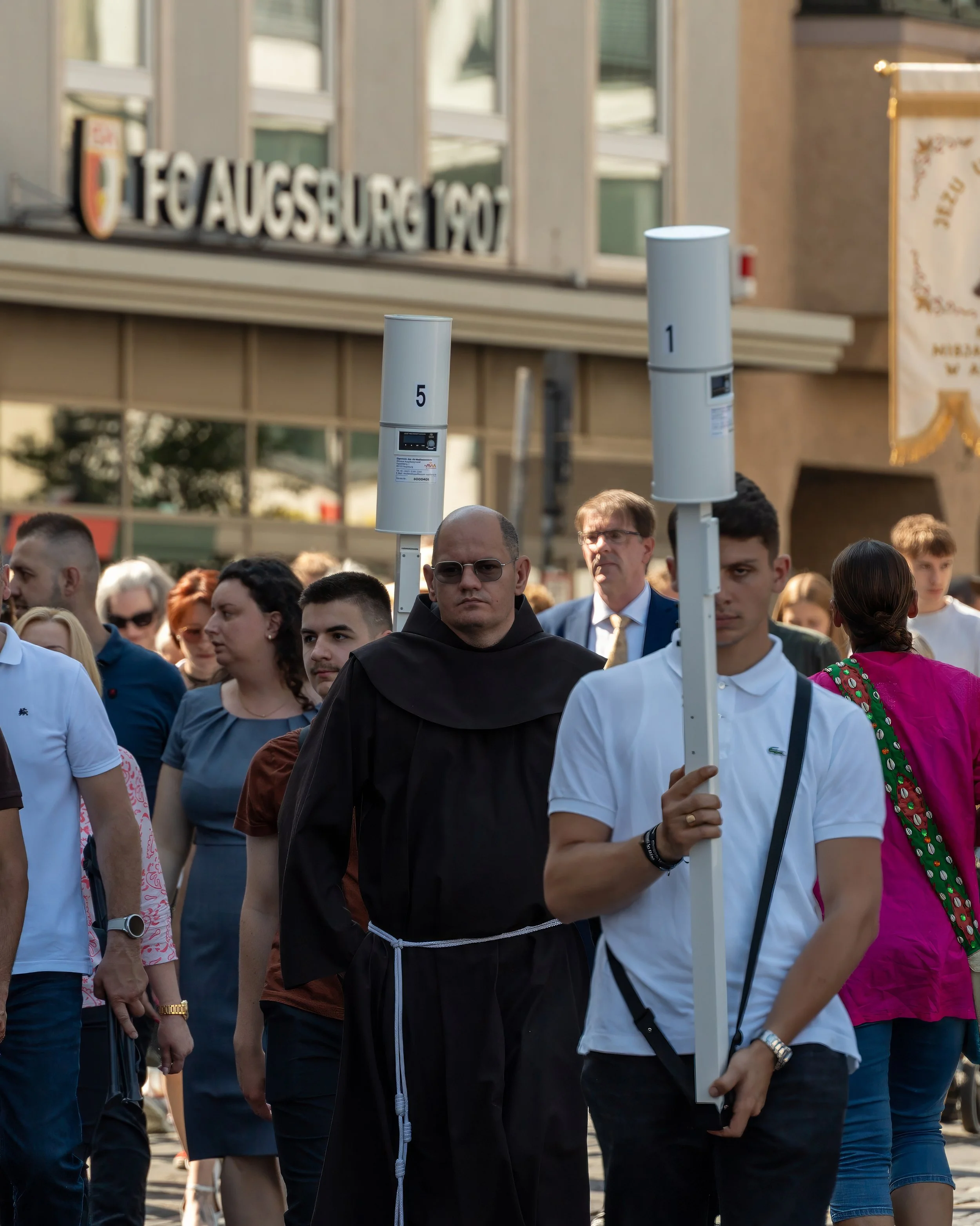 A group of people participating in a street procession, including a man dressed as a monk and a man holding a tall white pole with a speaker attached, during daytime.