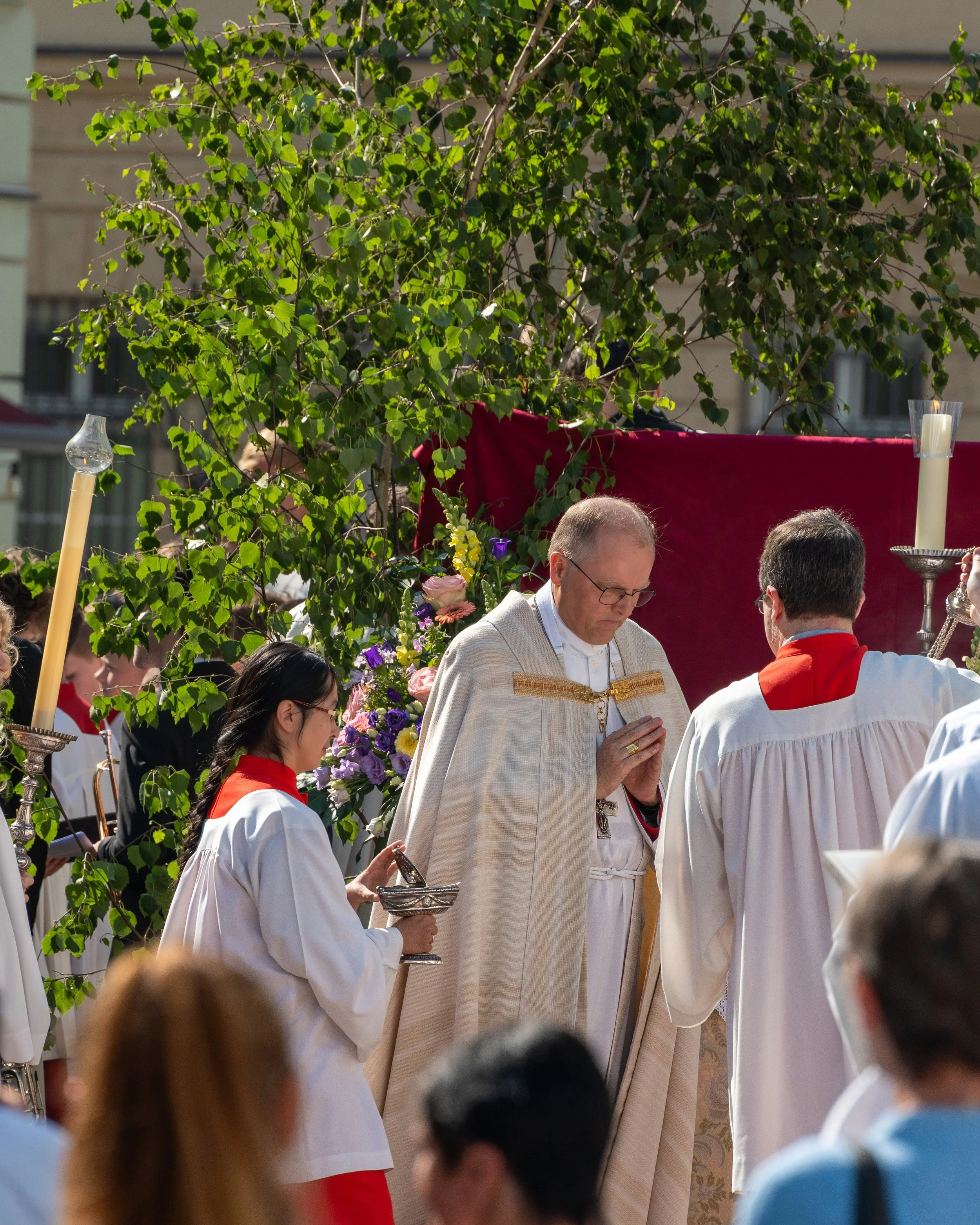 A religious outdoor ceremony with clergy and altar servers, surrounded by greenery and flowers.