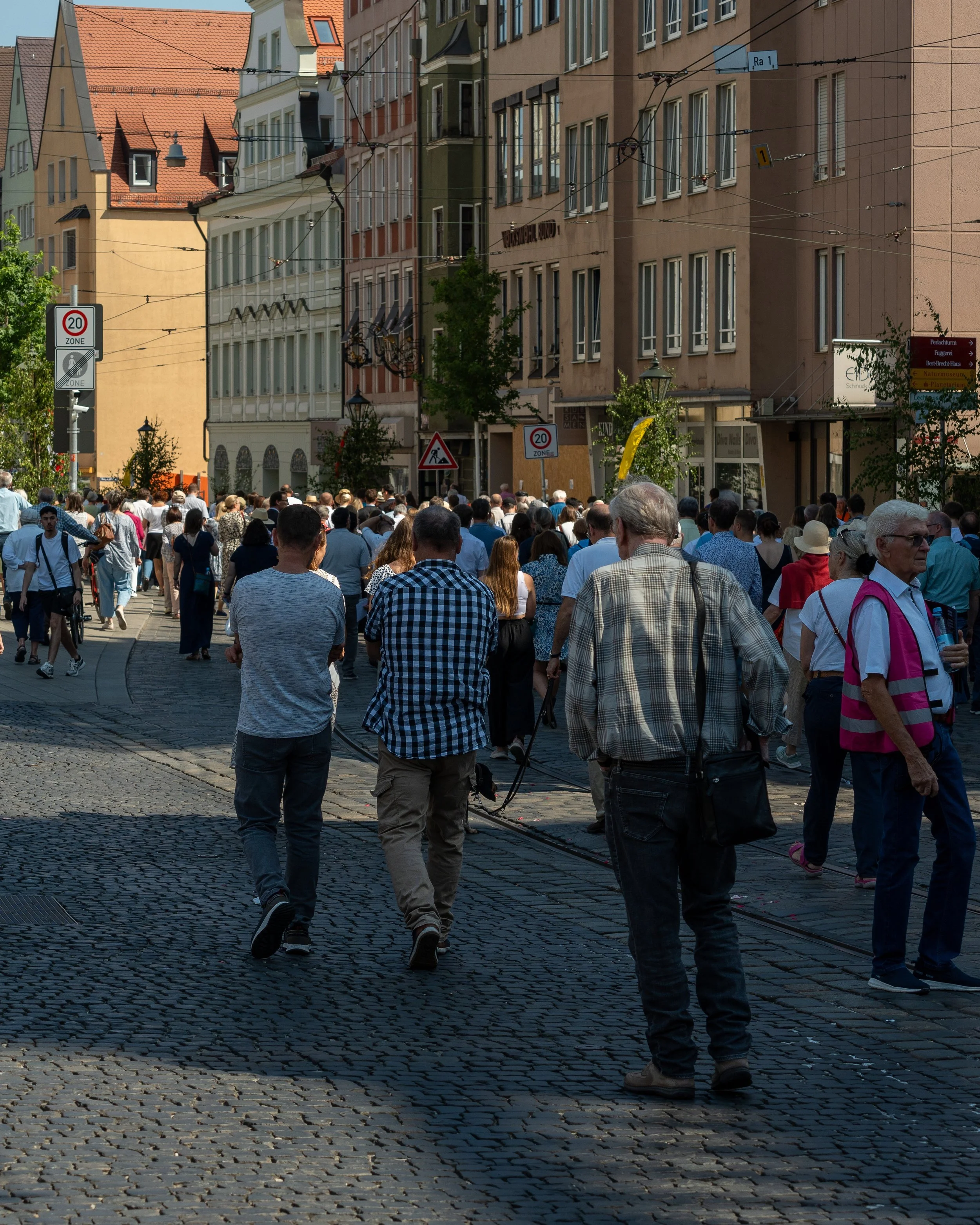 Crowd of people walking on a cobblestone street in an urban area with colorful buildings and overhead tram wires.
