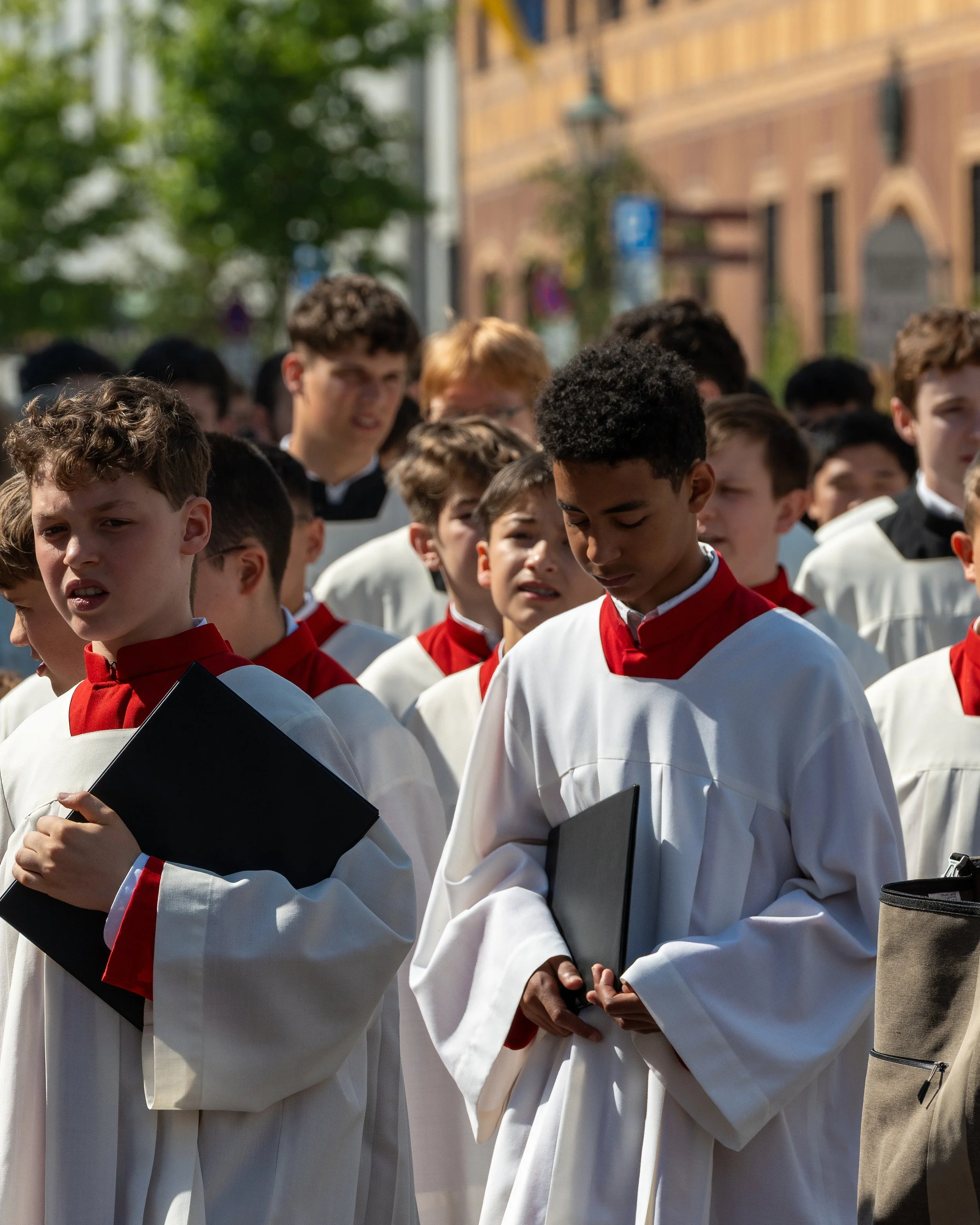 Group of young male students dressed in white choir robes with red collars, holding black folders, gathered outdoors during daytime for a ceremony or event.