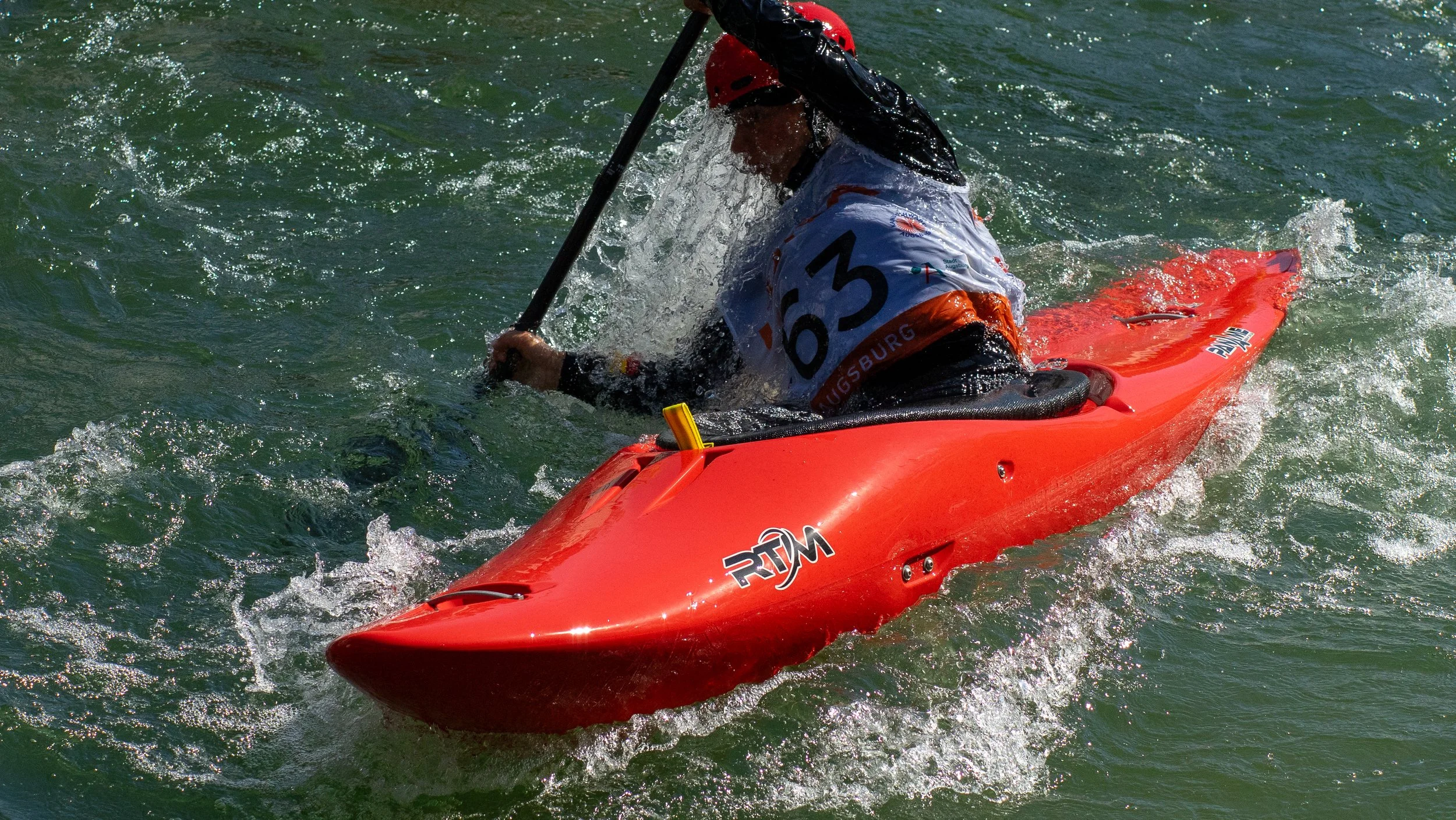 A person in a red kayak paddling in rough water, wearing a red helmet and a white jersey with the number 63.
