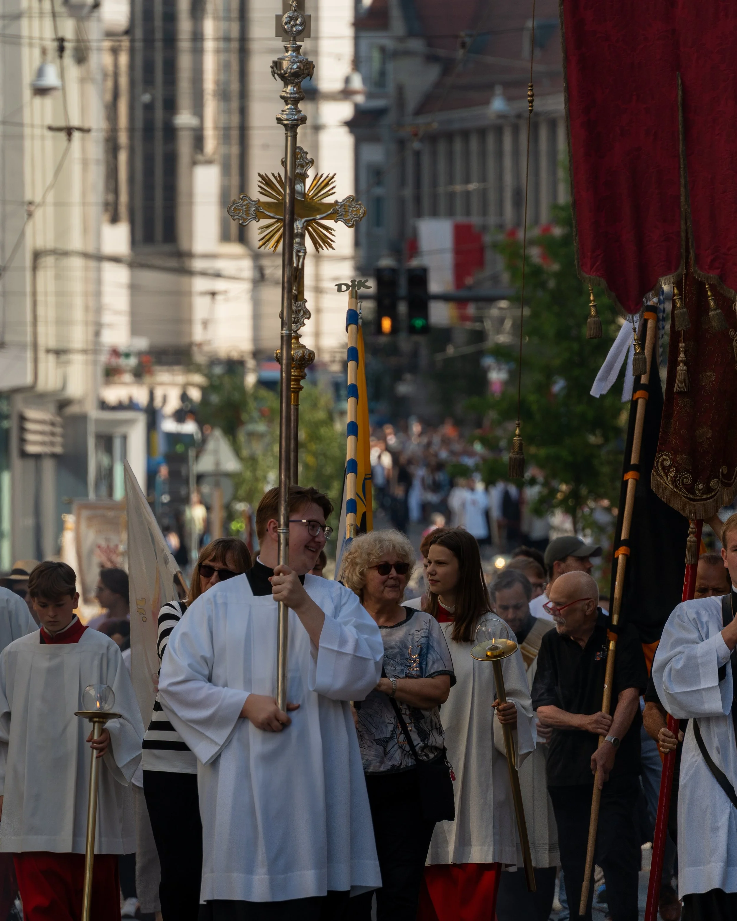 People participating in a religious procession, carrying crosses and candles, walking on a city street with buildings and a traffic light in the background.