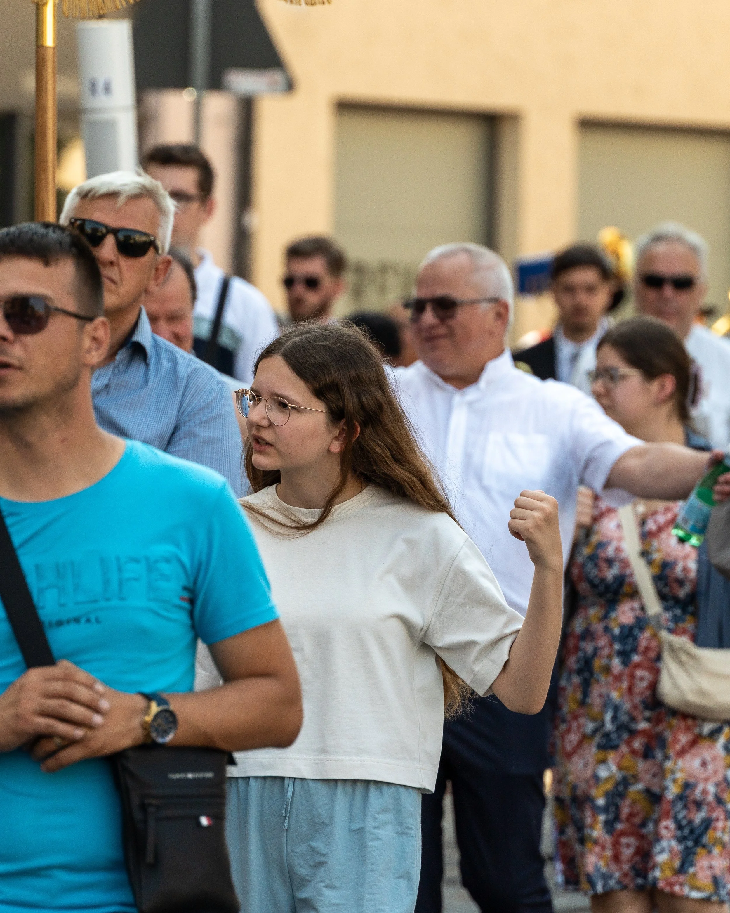 A diverse group of people, including young and older individuals, gathered outdoors, some wearing sunglasses, with a young woman in a white sweatshirt and glasses in the foreground showing her flexed arm.