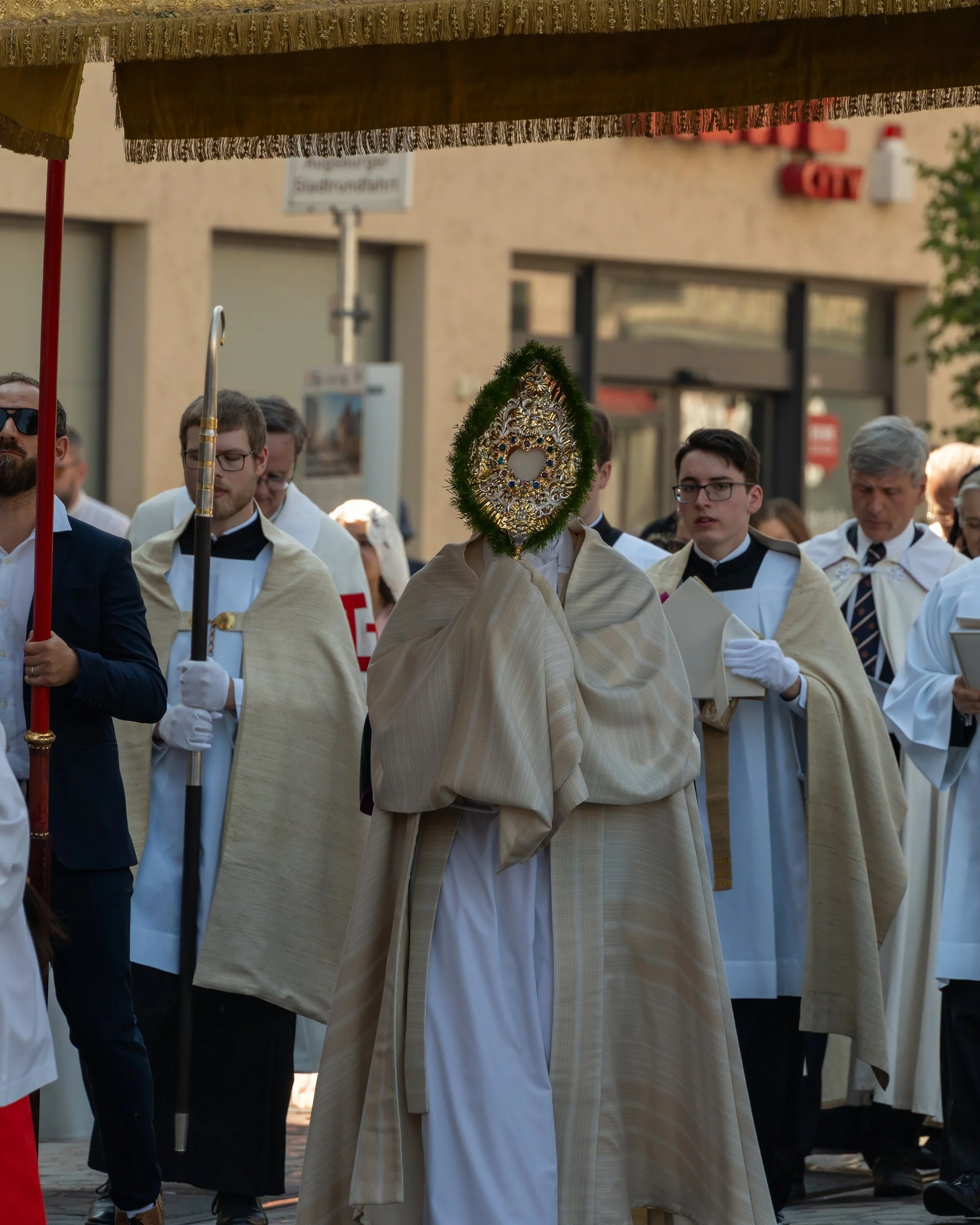 A religious procession with several men dressed in ceremonial robes, one holding a staff, and another holding a book, walking together on a city street. The central figure's face is obscured by an ornate decorative item, possibly a religious relic or