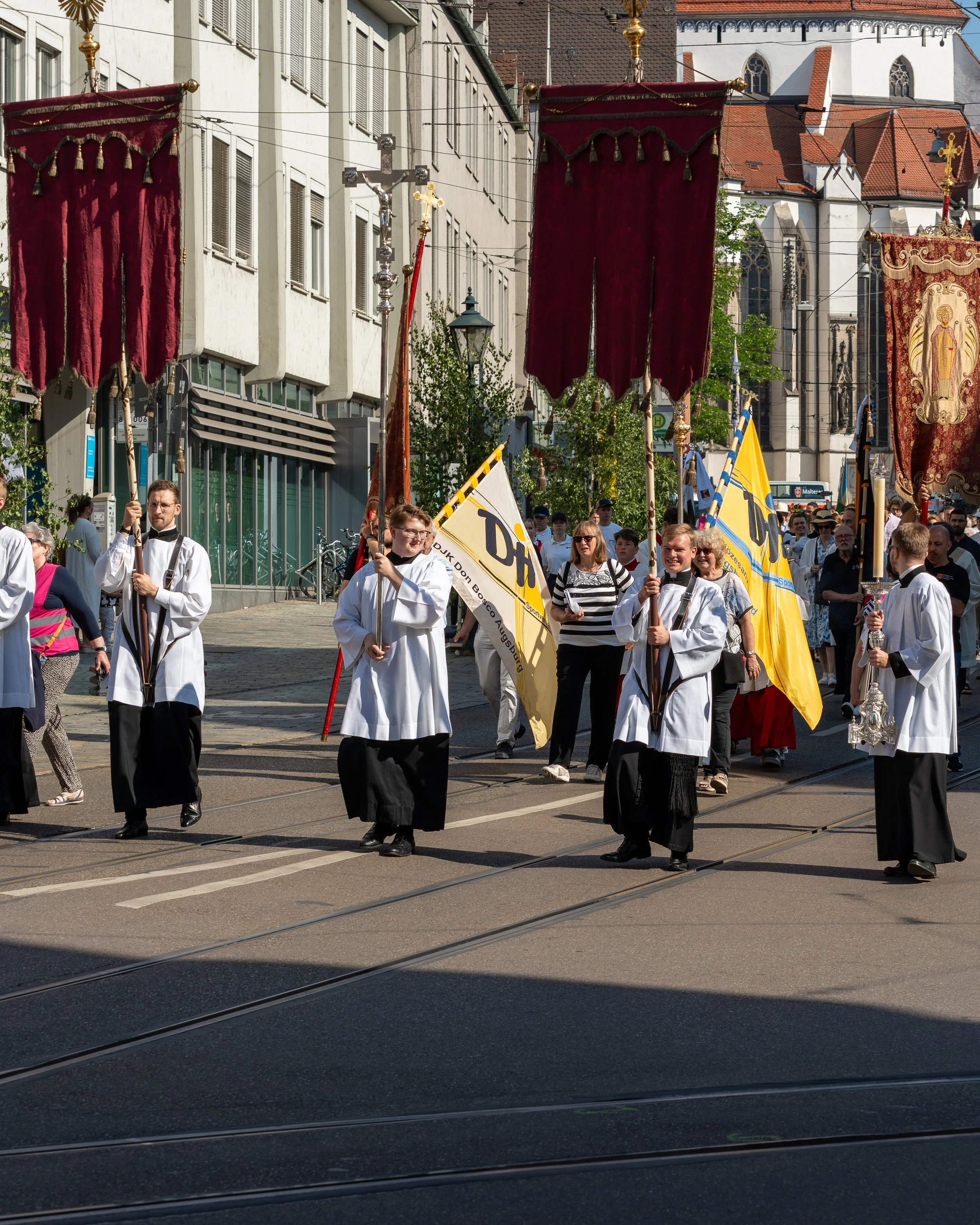 People participating in a religious procession carrying religious banners and flags, walking on city streets with onlookers in the background.