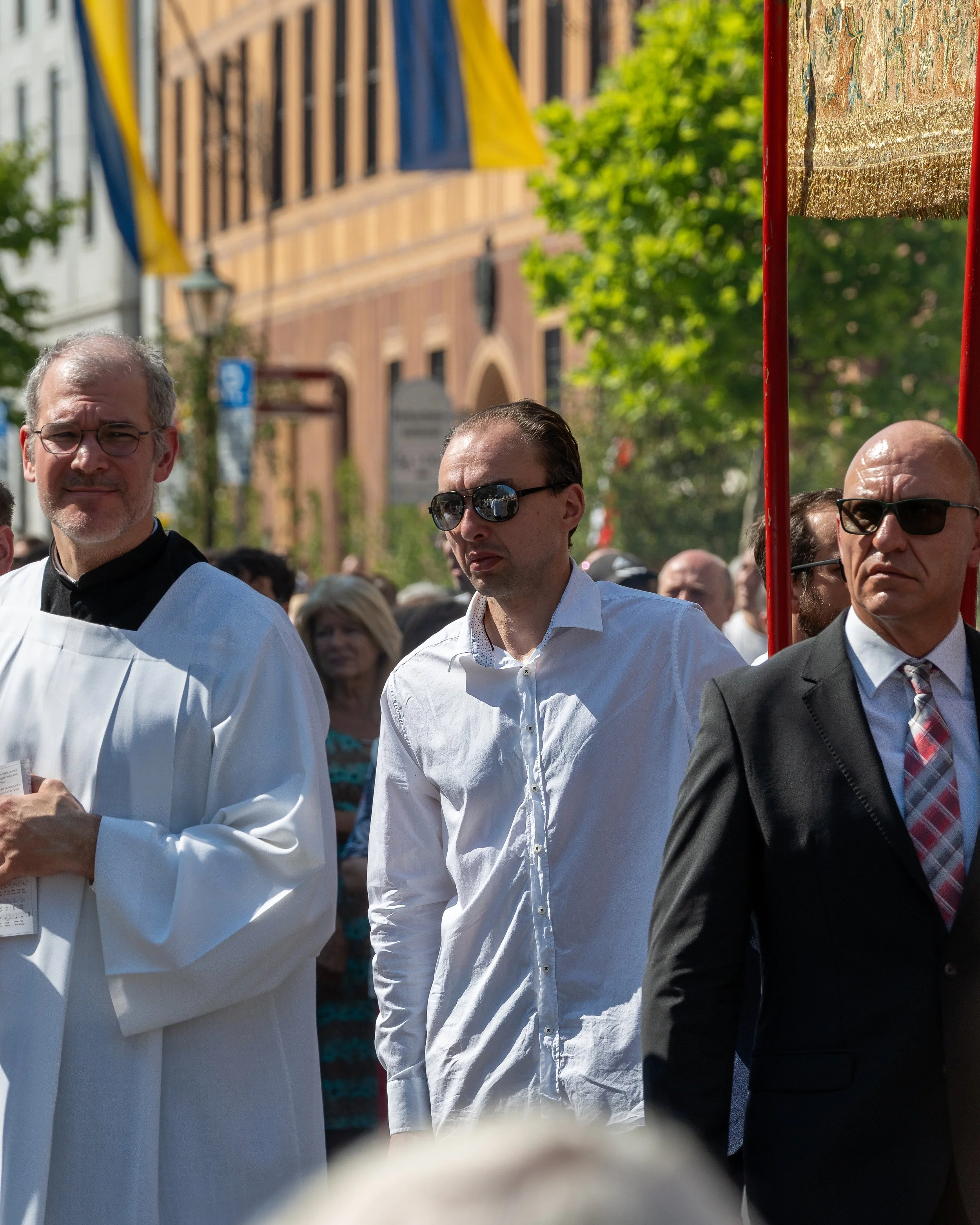 A group of men participating in a formal outdoor procession or event, some wearing sunglasses and formal attire, standing among a crowd with a historic building and flags in the background.