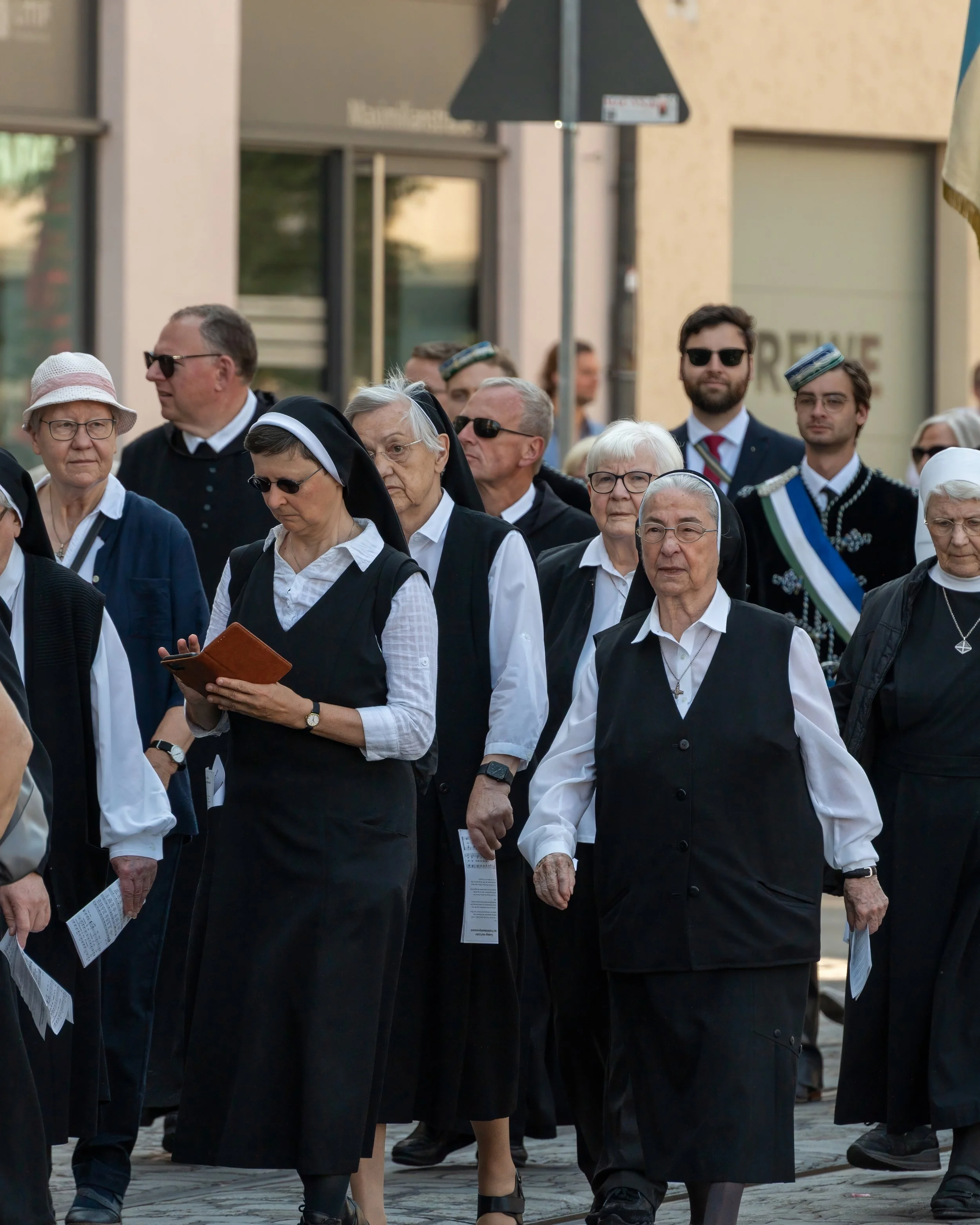 A group of nuns, priests, and other individuals participating in a procession or march outside, some dressed in religious habits and others in formal attire, with buildings and a street in the background.