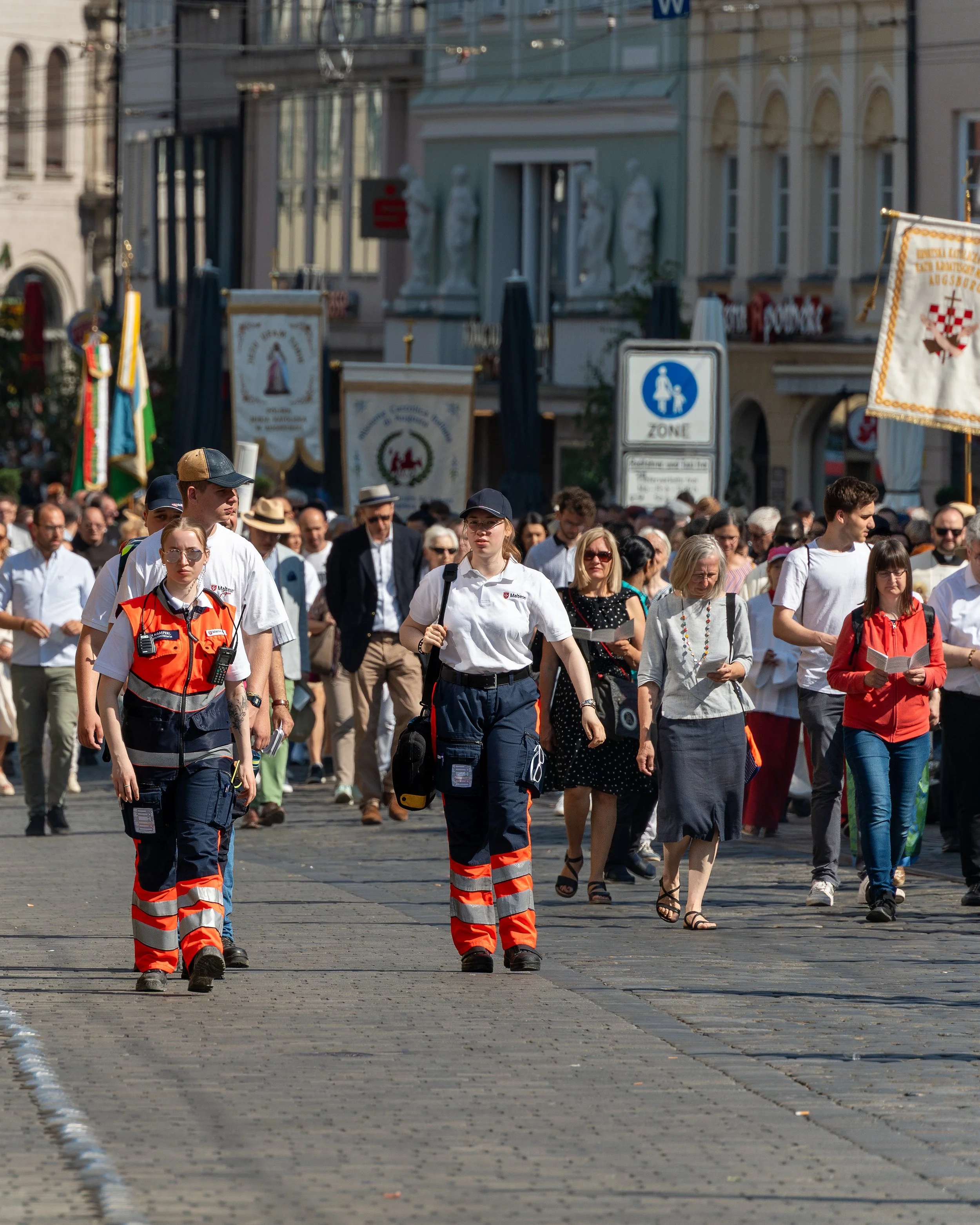 A group of people participating in a procession or march, walking along a city street with historic buildings in the background. Some participants are reading from booklets, and two emergency responders are leading in the front.
