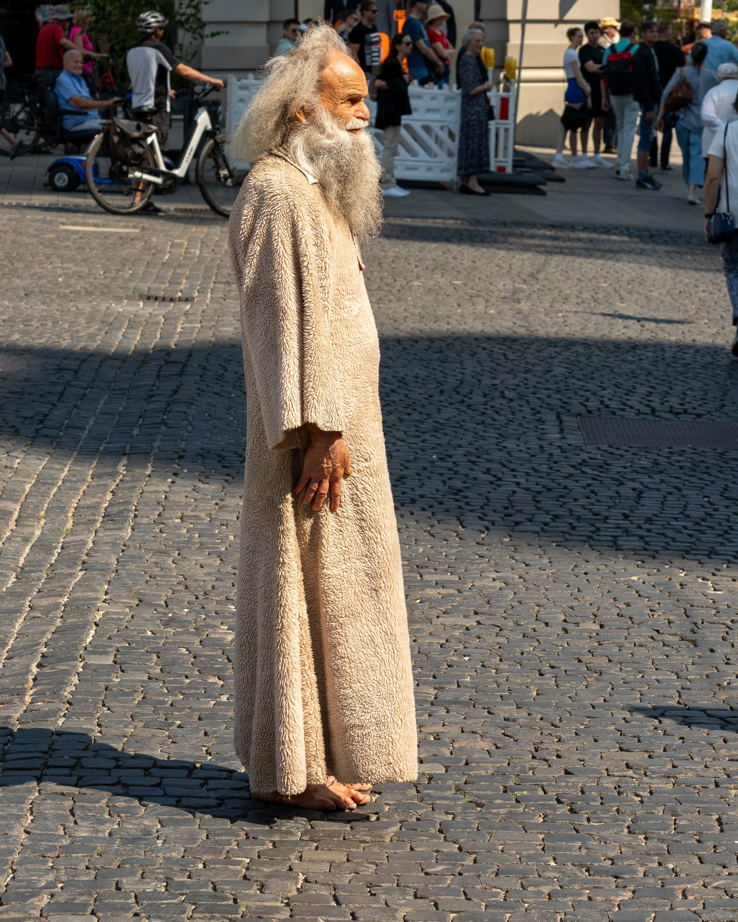 An elderly man with long white hair and beard, wearing a beige, fuzzy coat and barefoot, standing on cobblestone street with shadow cast behind him and a crowd of people in the background.