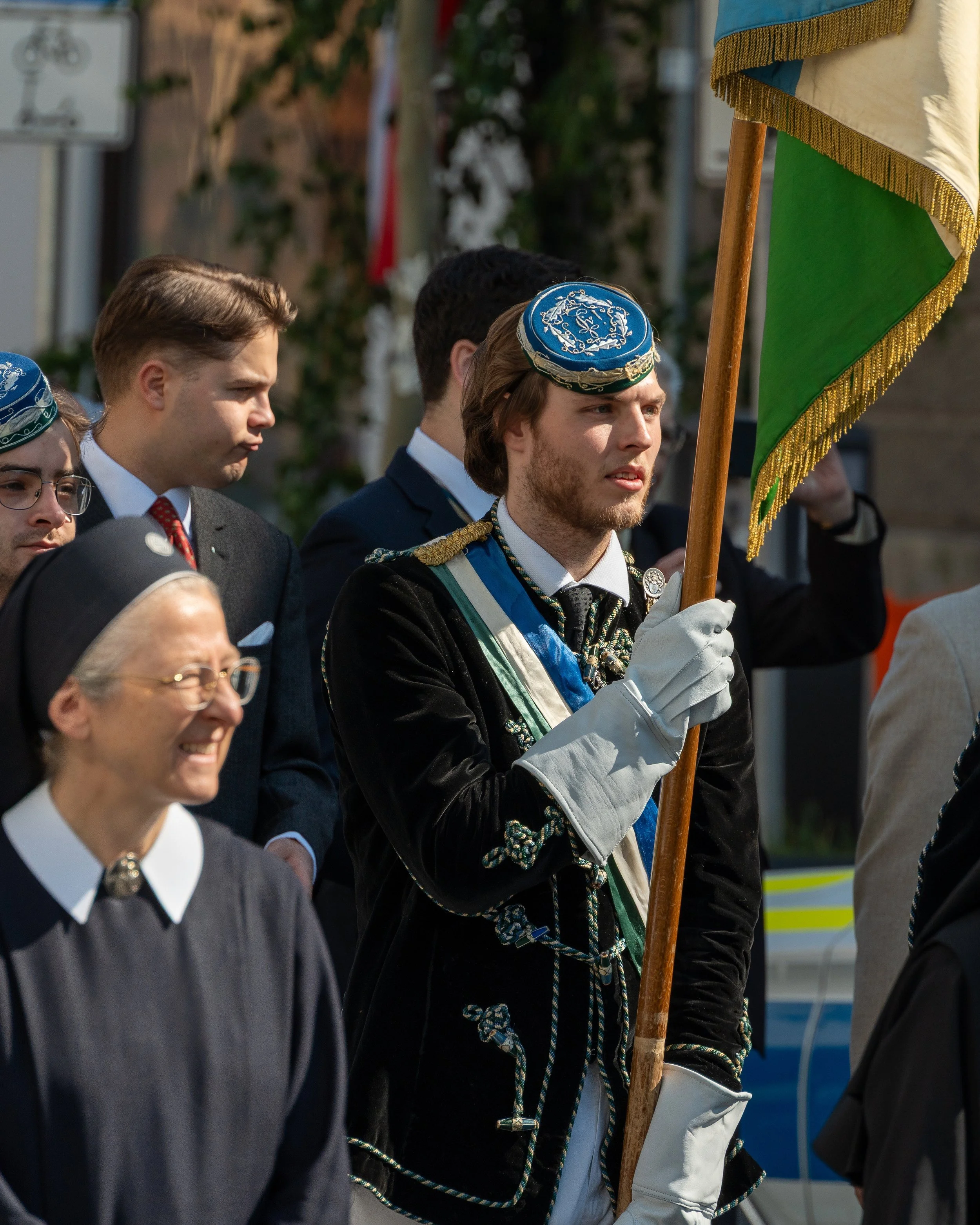 A young man dressed in ceremonial attire holding a flag during a parade or ceremonial event, surrounded by others including a nun, in an outdoor setting.