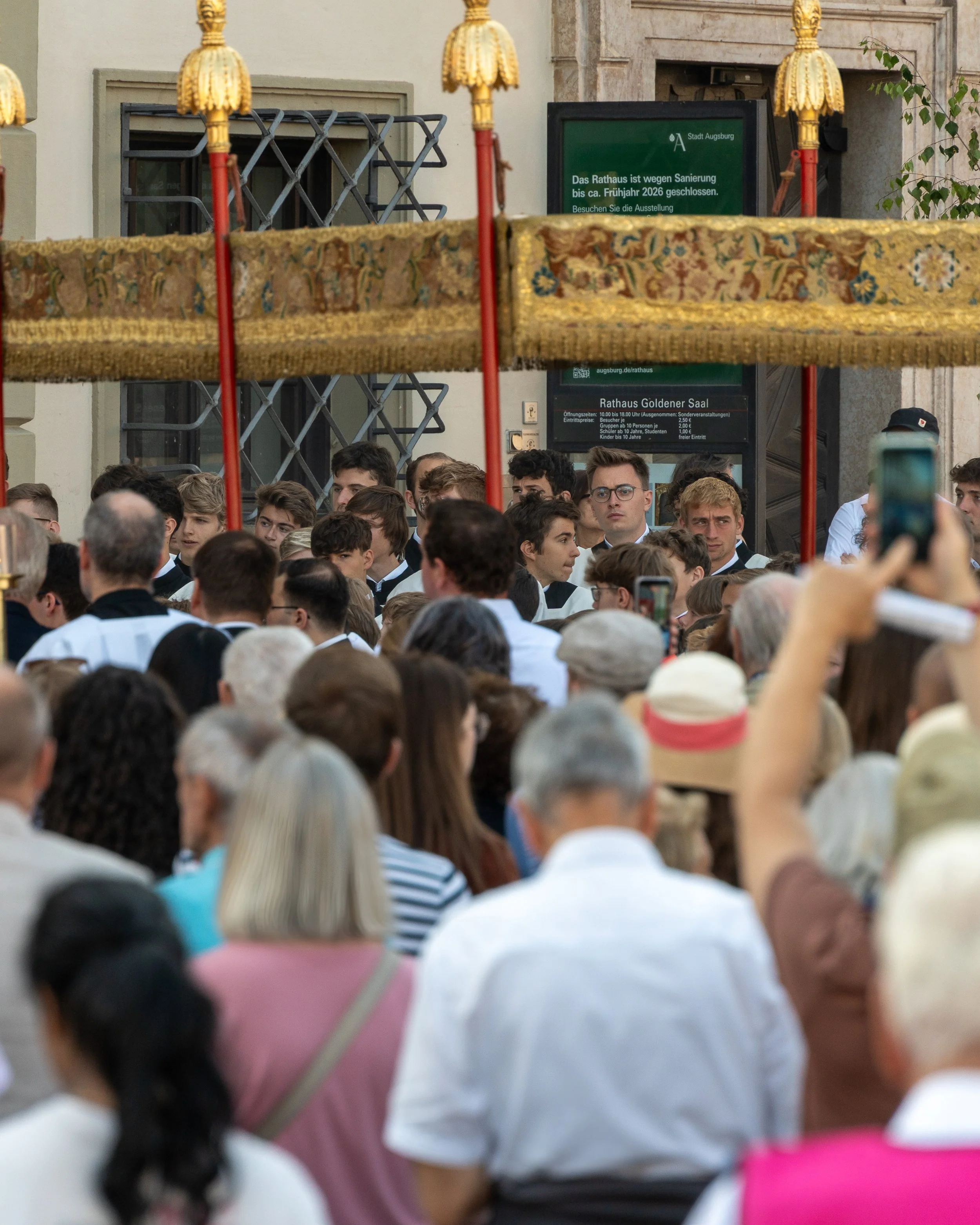 A crowd of people gathered for a public event or ceremony, with a group of young men in formal attire standing behind a canopy, and some people in the crowd taking photos.