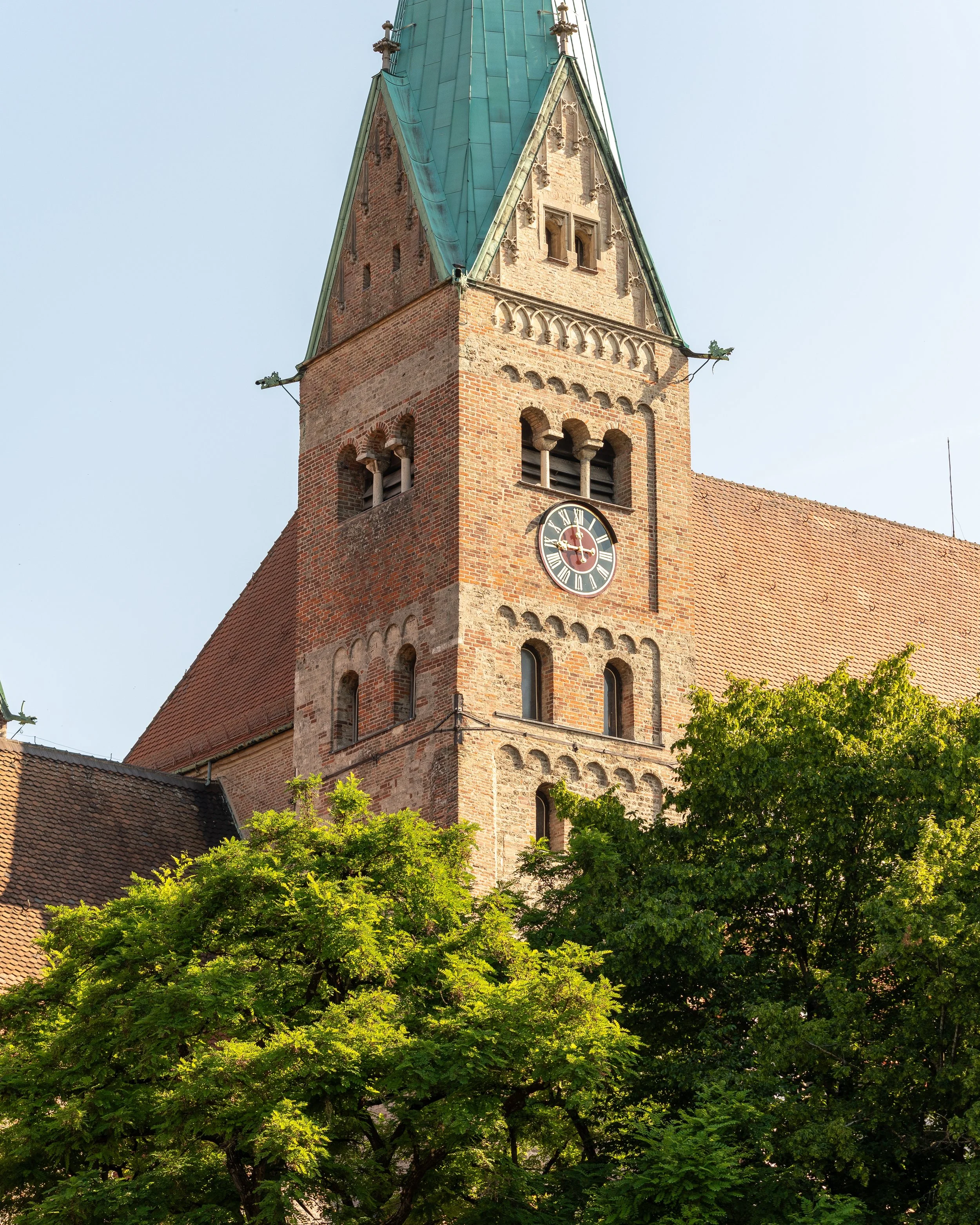 A historic brick clock tower with a green pointed roof, surrounded by green trees, under a clear blue sky.