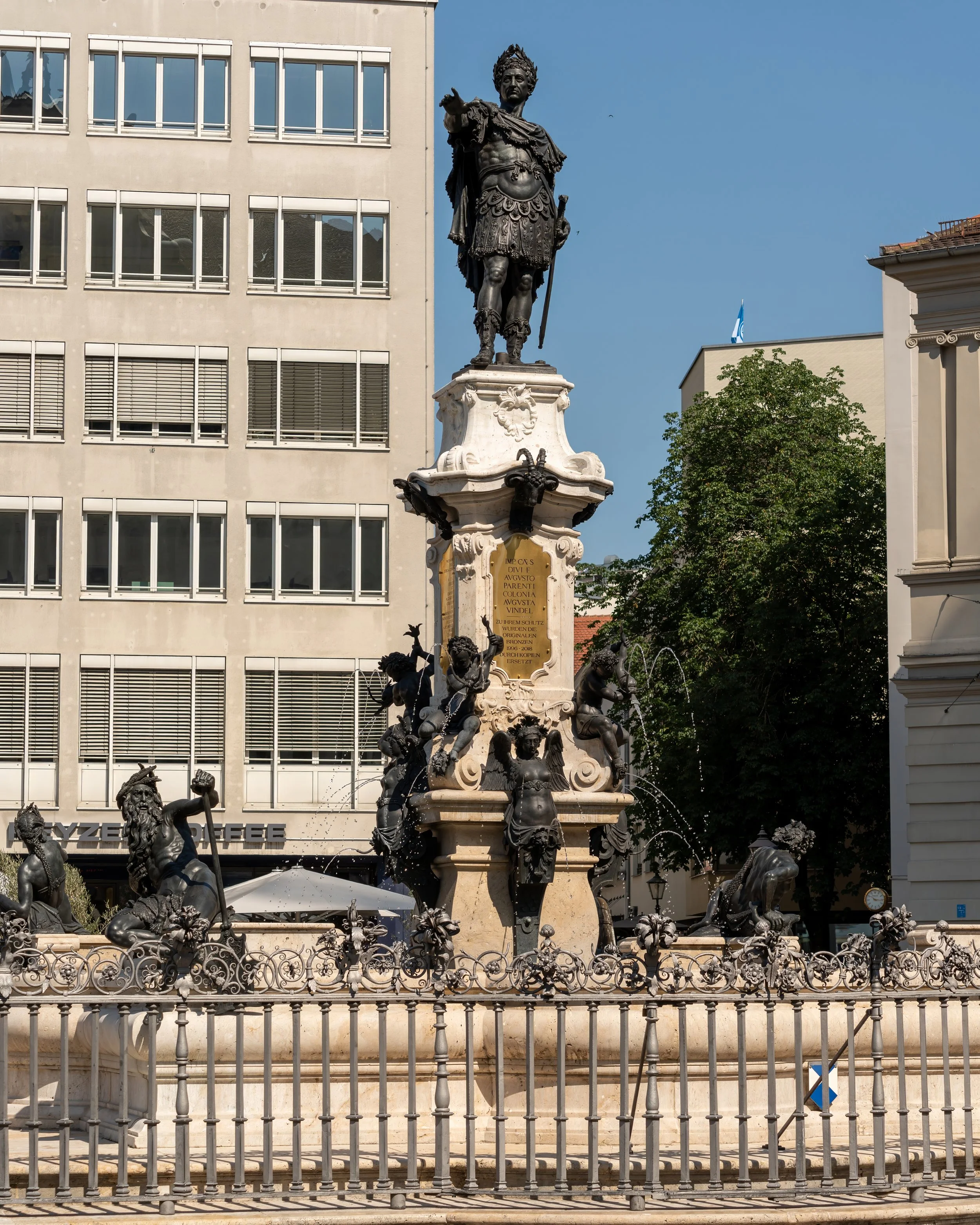 A tall, ornate fountain with sculptures of mythological figures and a statue of a man in armor at the top, set against modern buildings and trees.
