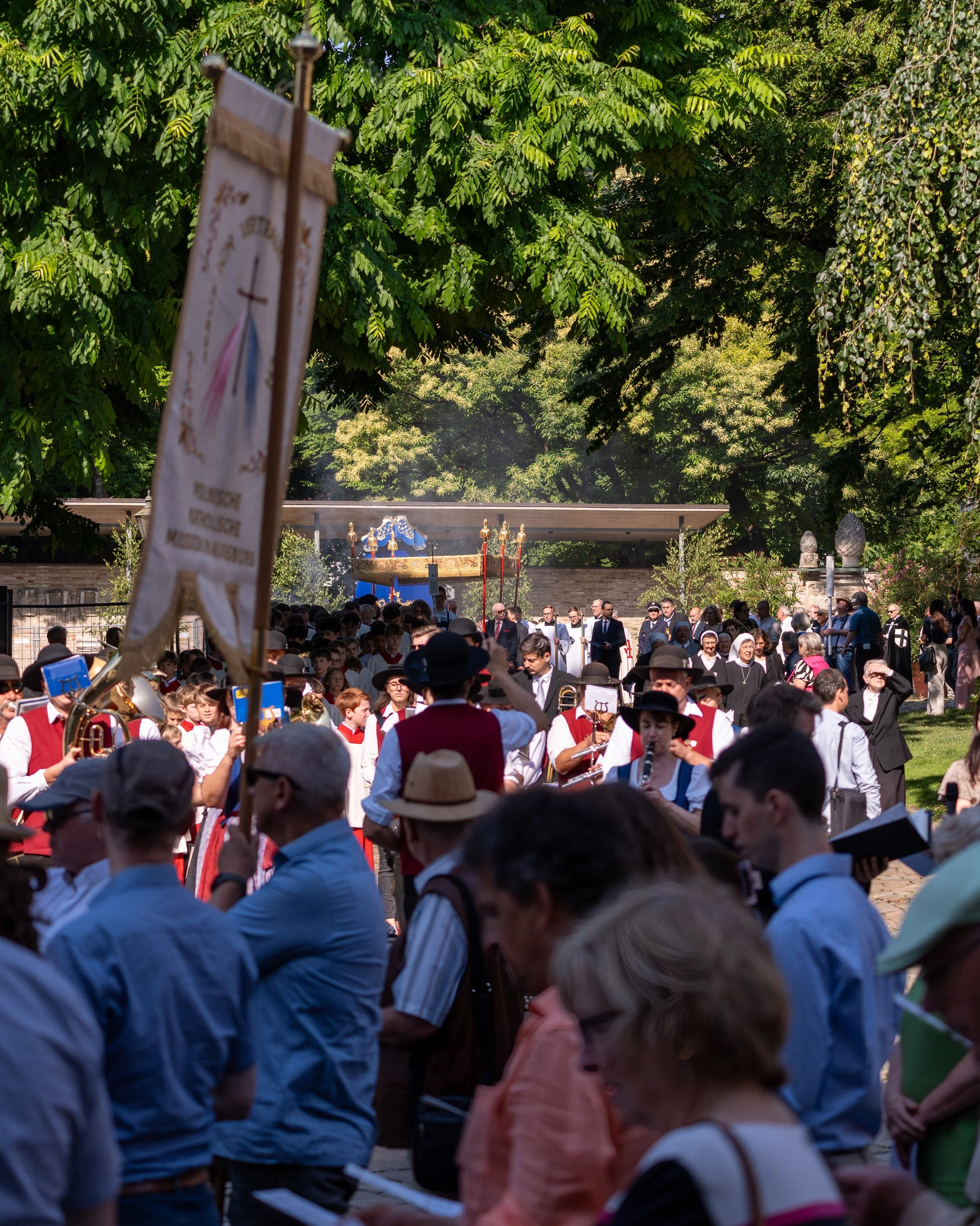 A large outdoor gathering of people, including musicians in traditional attire playing instruments, with a ceremonial procession and a decorated float in the background. Green trees surround the scene on a sunny day.