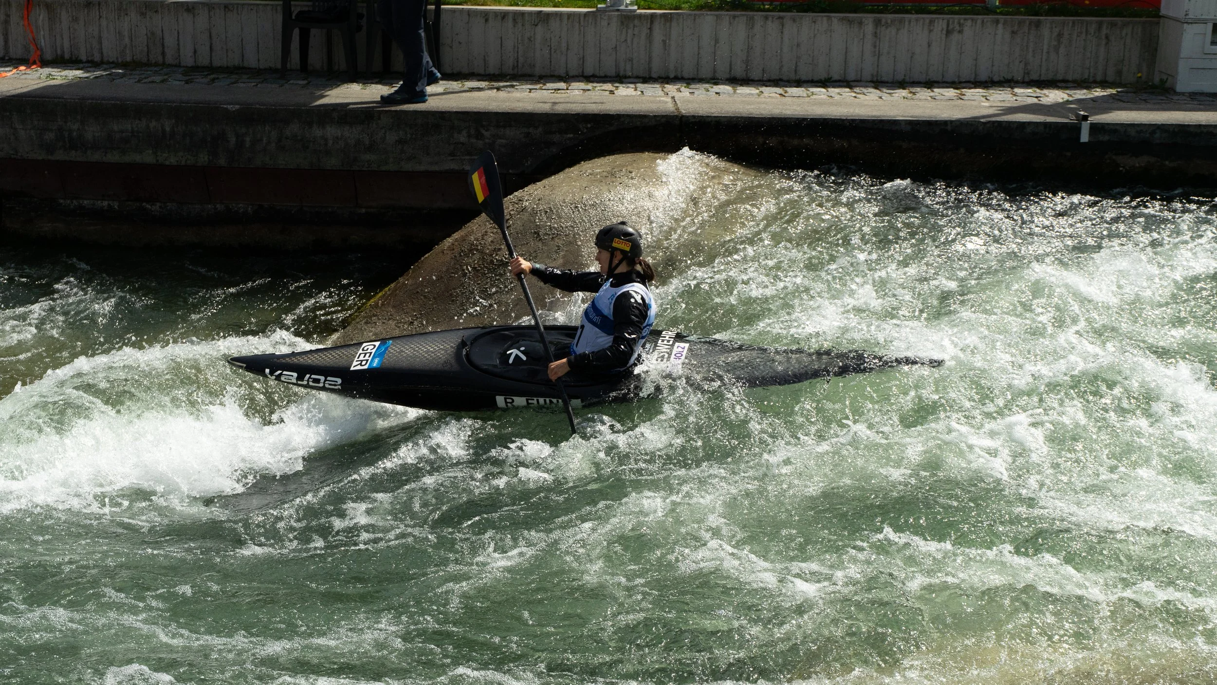 A person kayaking in turbulent water near a dock, wearing a helmet and a life jacket, holding a paddle with a flag design.