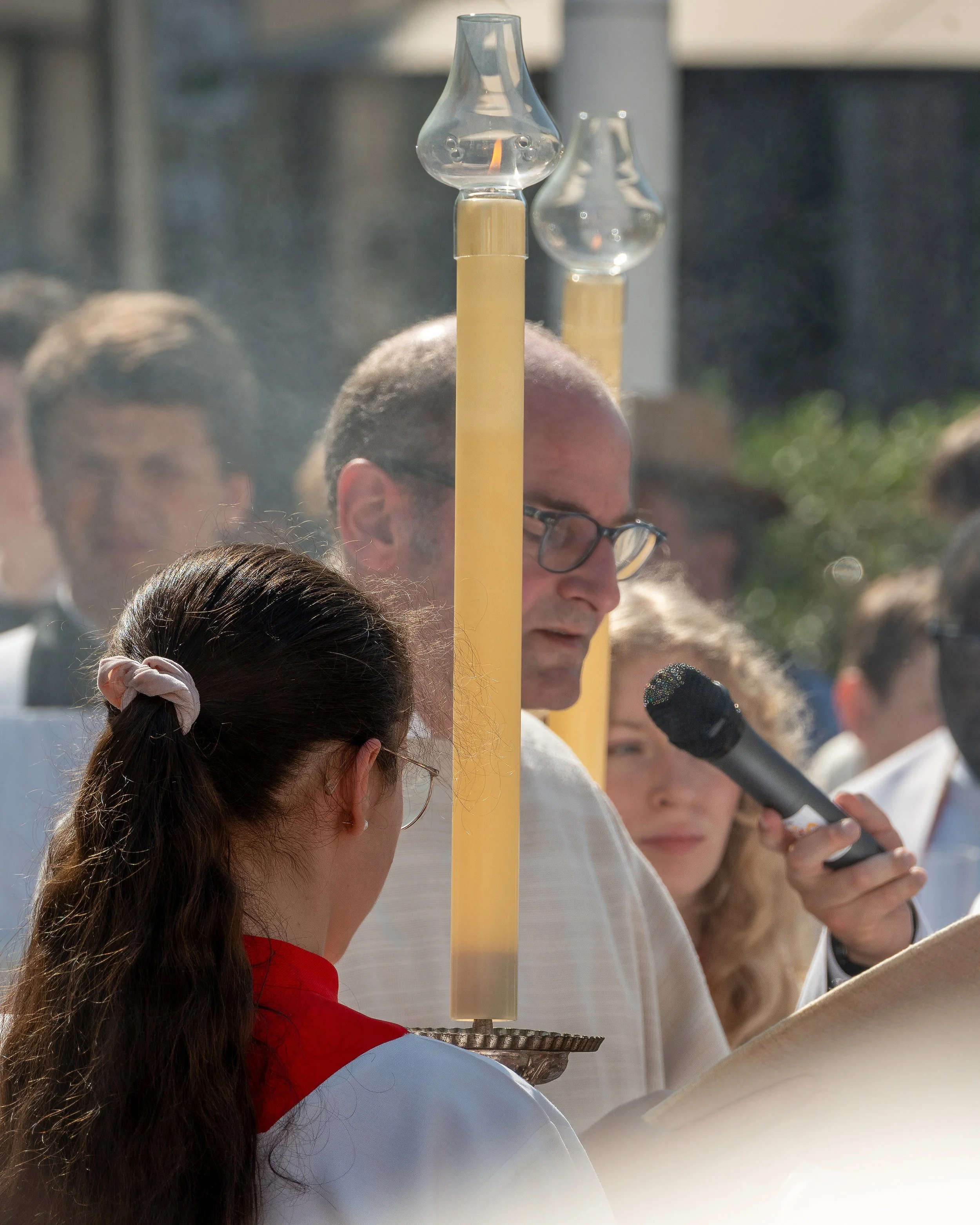 A religious outdoor ceremony with a man speaking into a microphone, standing behind a candle with glass chimneys, during the daytime.