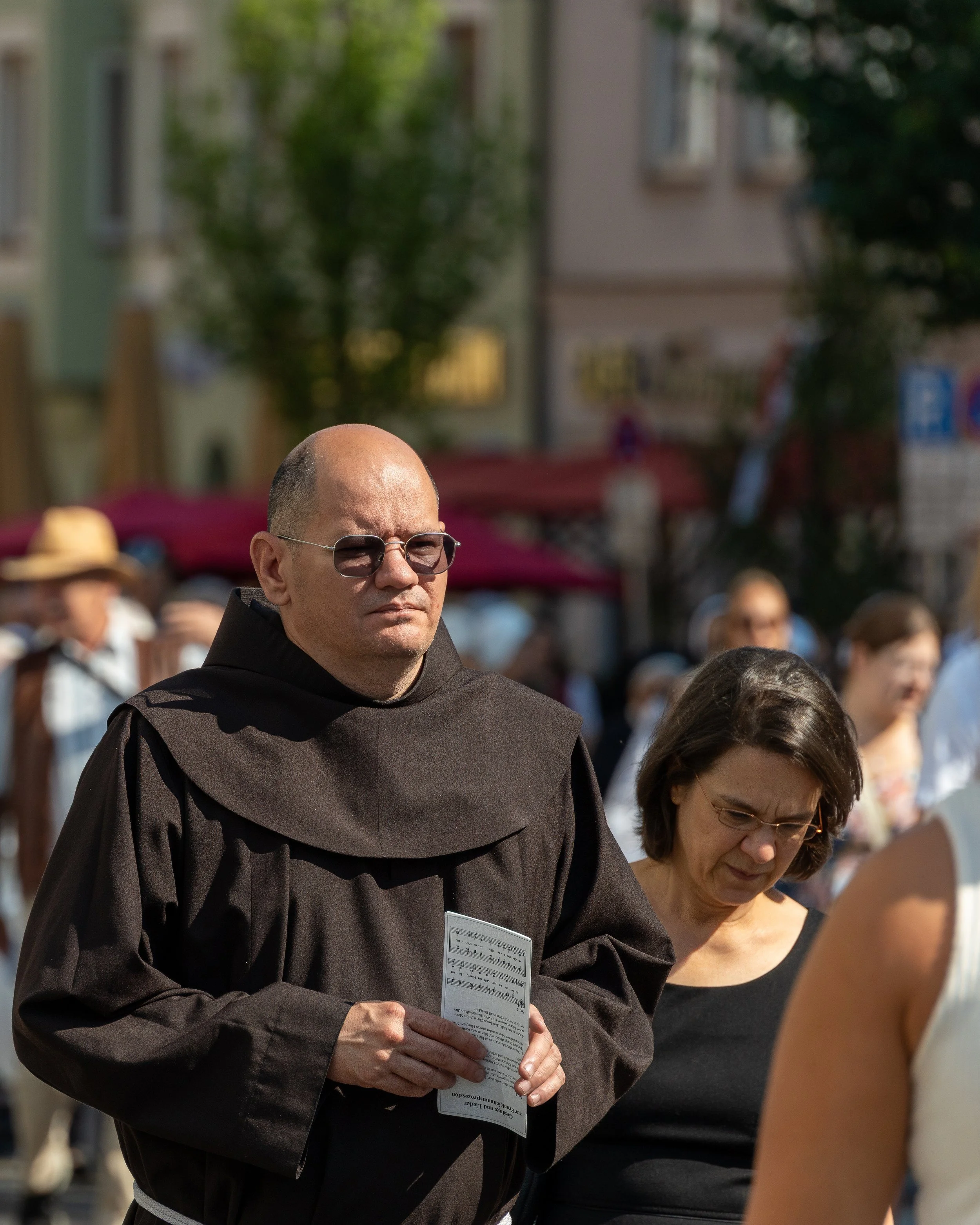 A man dressed as a monk holding a pamphlet, standing in a crowd outdoors during the day.