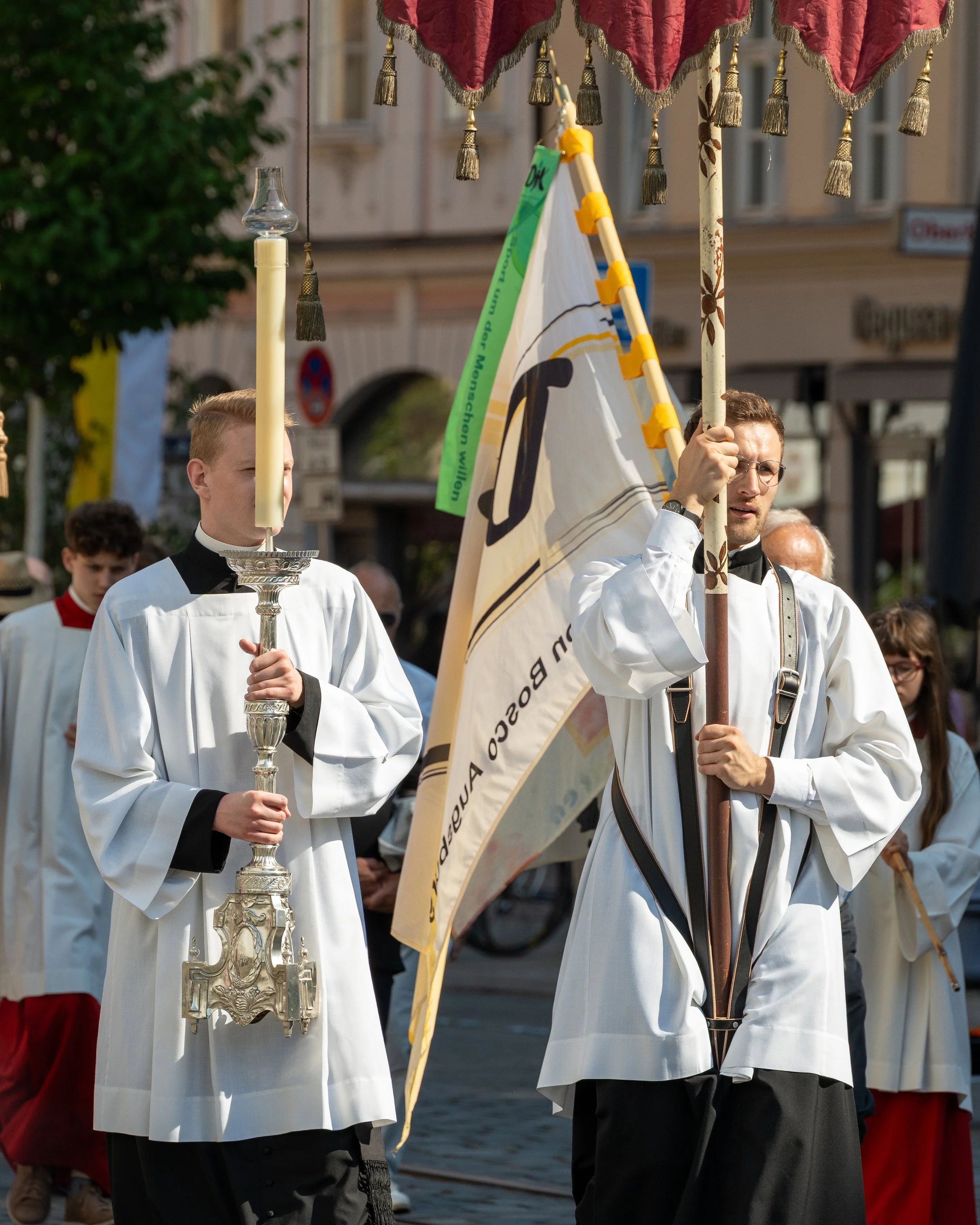 Two young men dressed in white robes participating in a religious procession. One holds a large silver candlestick with a tall candle, the other carries a flag with a wooden pole. People in similar attire are visible in the background.