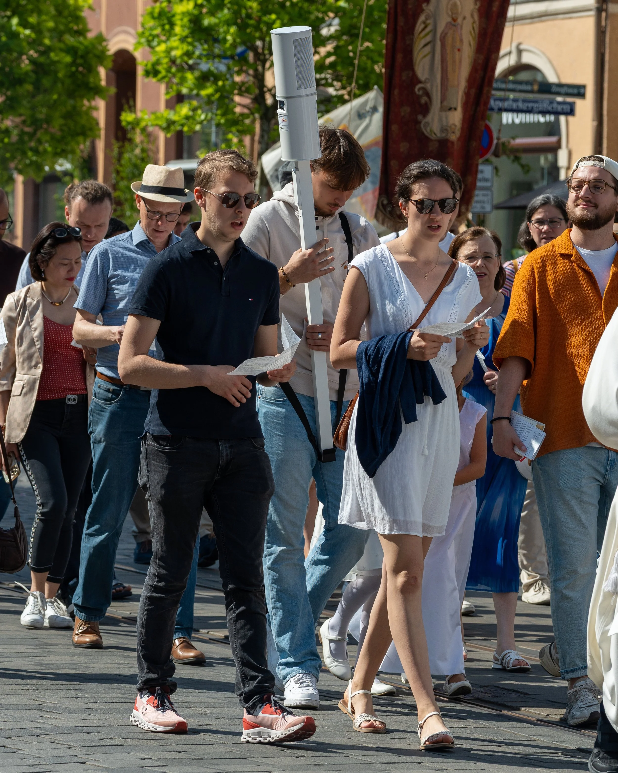 A group of people walking outdoors, some holding pamphlets, with trees and buildings in the background.