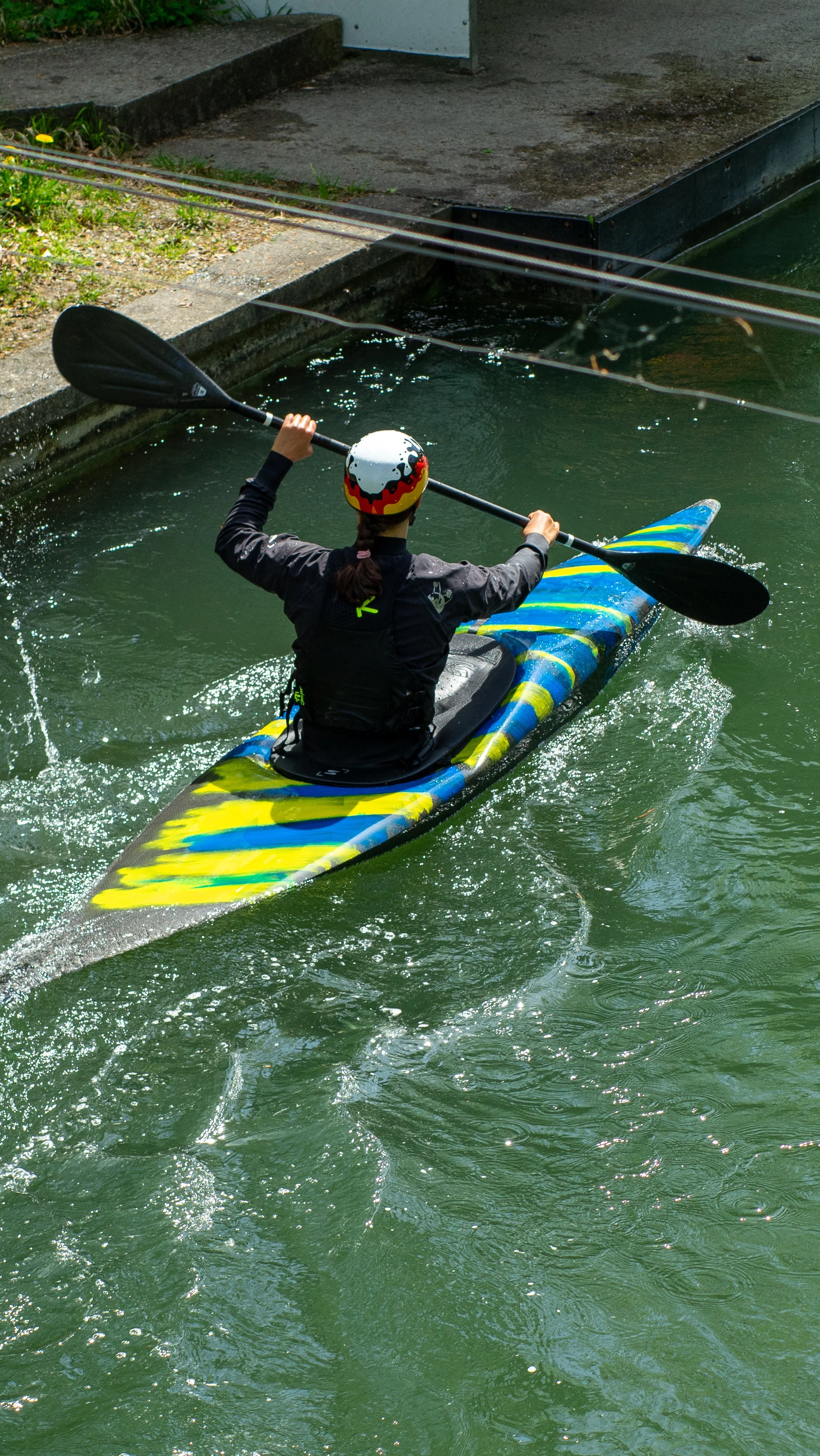 Person kayaking in a narrow waterway, wearing a colorful helmet and black jacket, paddling with a double-bladed paddle.