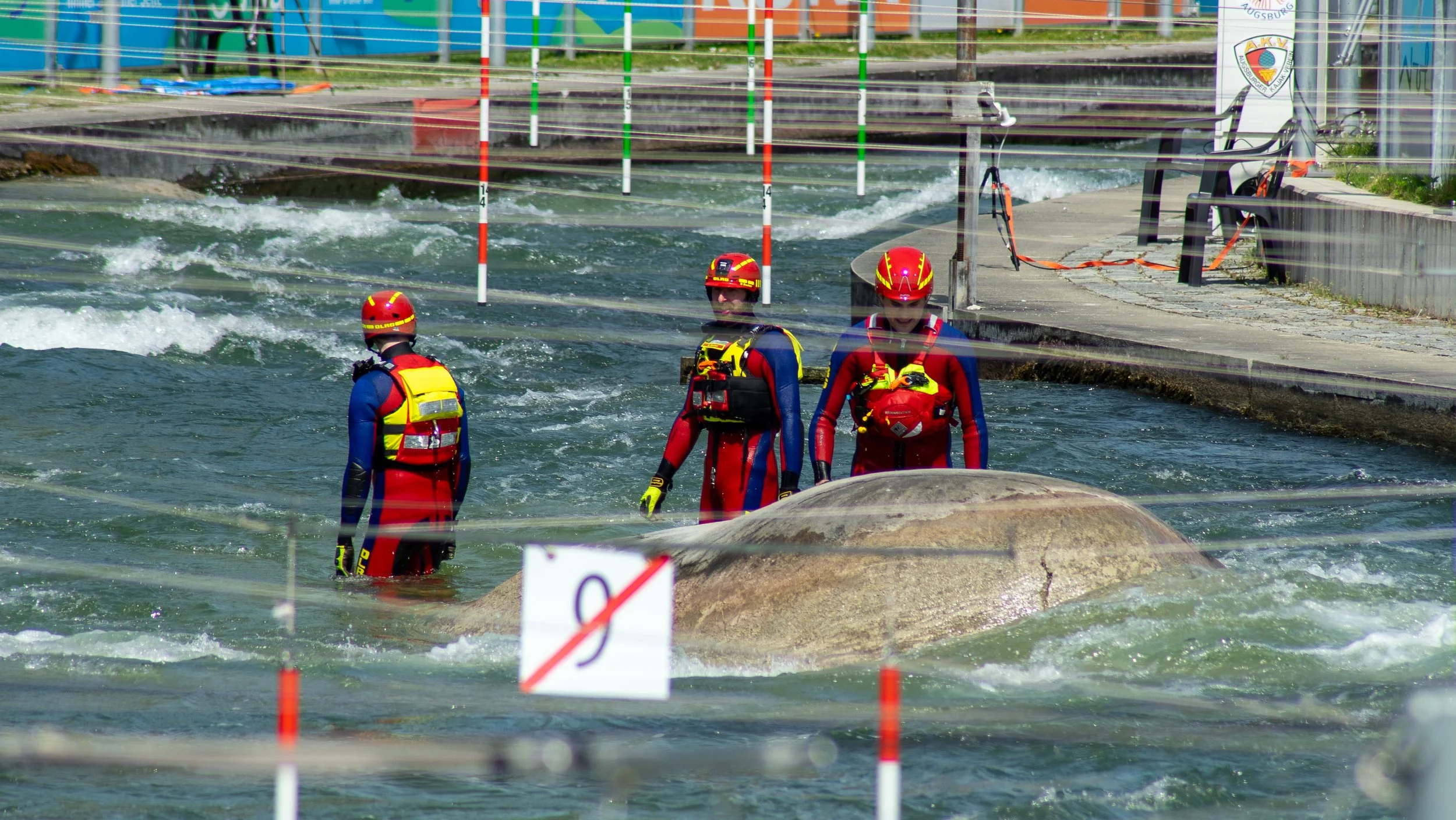 Three rescue workers in red and blue wetsuits and red helmets wading through a water channel, possibly during training or rescue operation, with barriers and equipment along the banks.