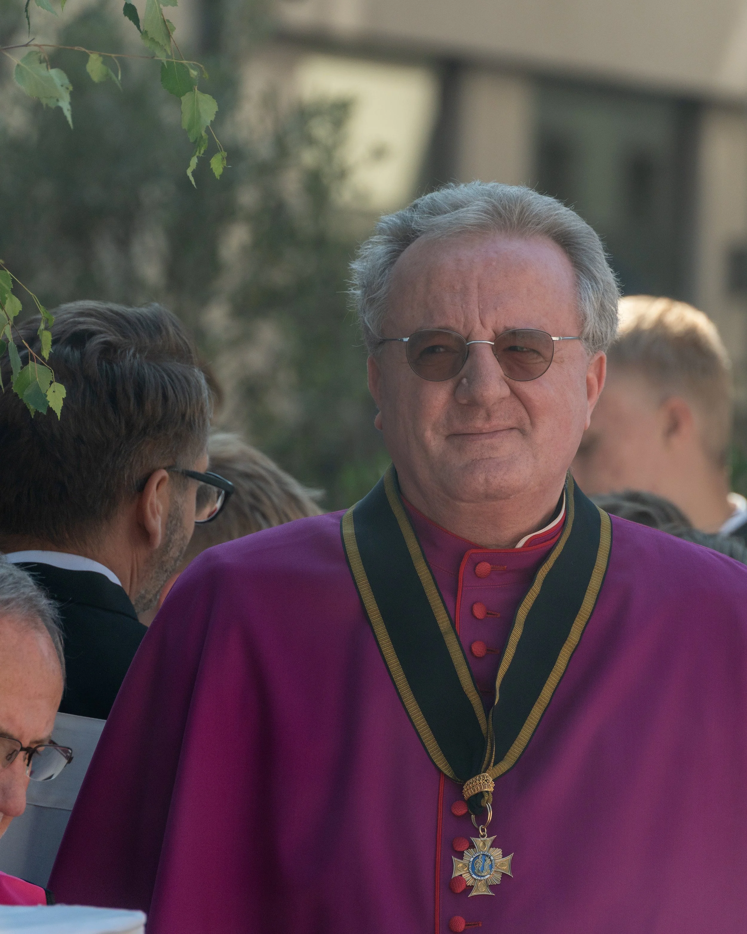 A man wearing a purple robe with gold trim, sunglasses, and a medal hanging from a ribbon around his neck, standing among other people outdoors.