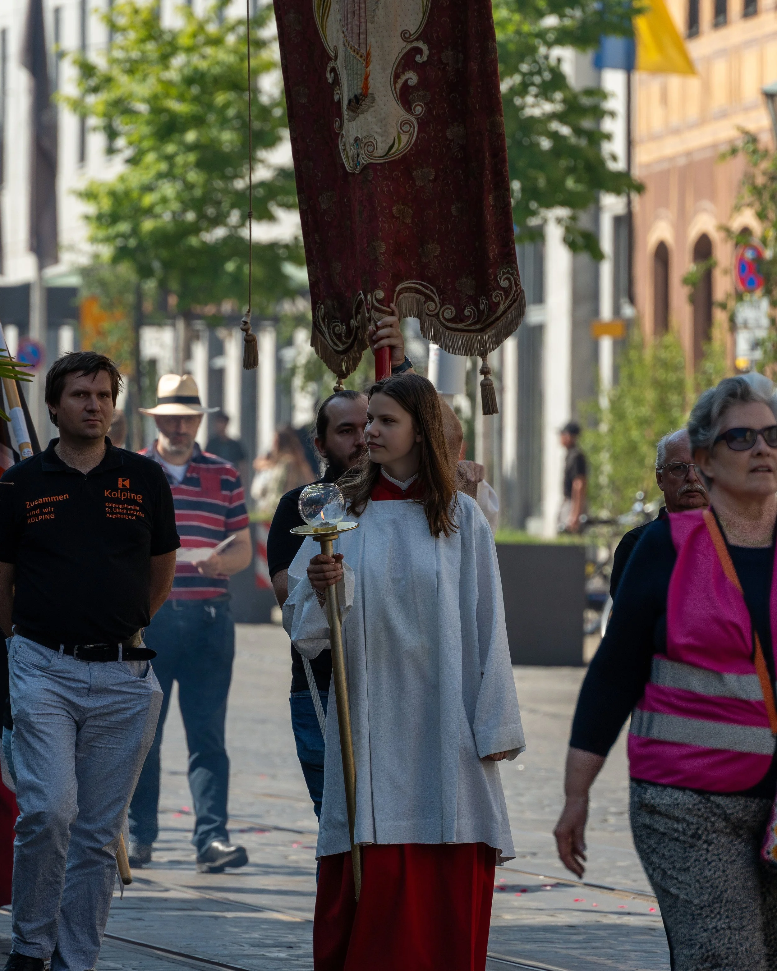 A young woman in a white religious robe holds a shiny orb and a staff while standing in a street parade. She is surrounded by other parade participants, some of whom are dressed casually. The woman is located slightly to the right and is holding a la