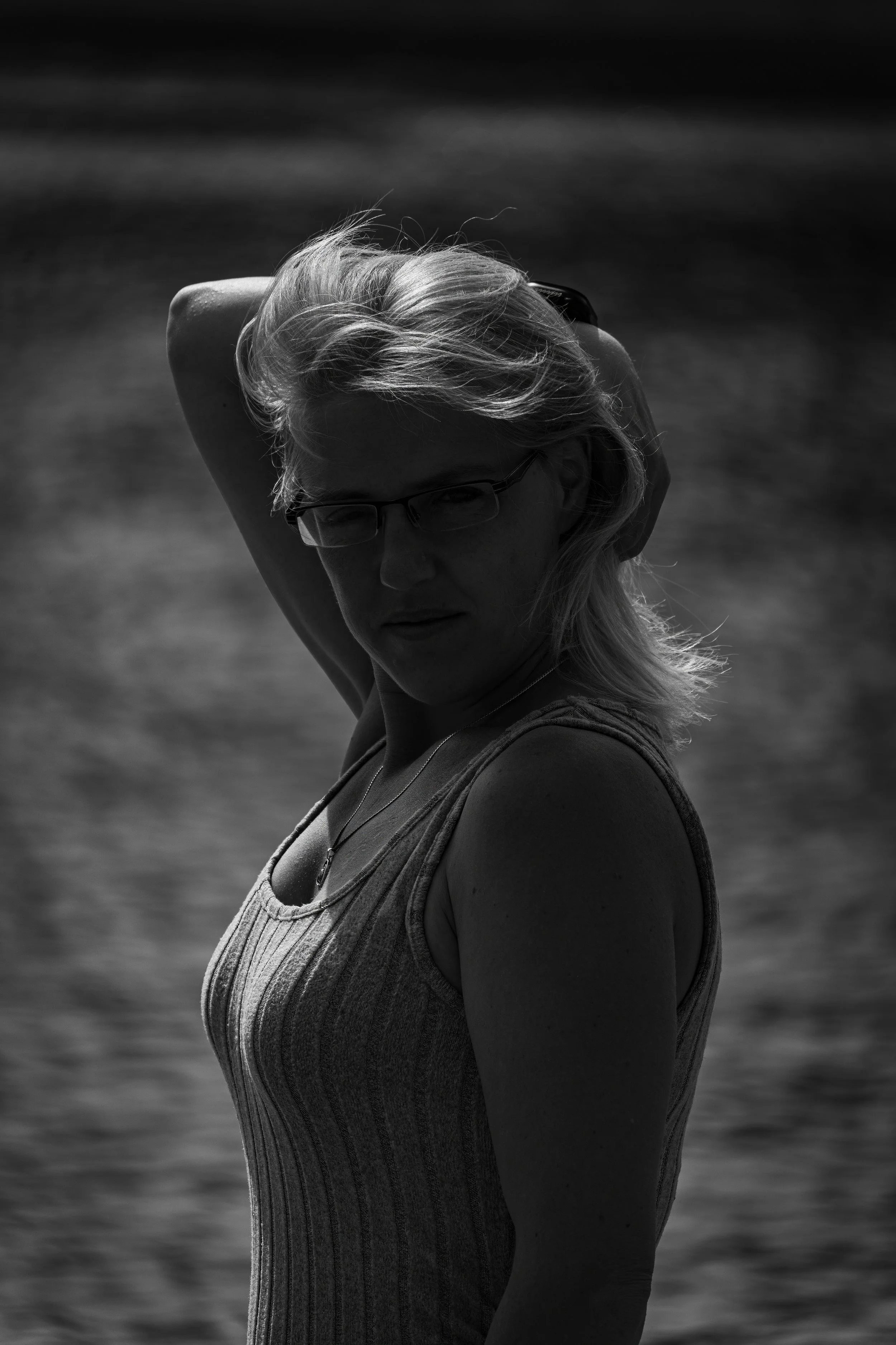 Black and white photo of a woman with glasses, wearing a striped tank top, standing outdoors near a body of water with the horizon in the background.