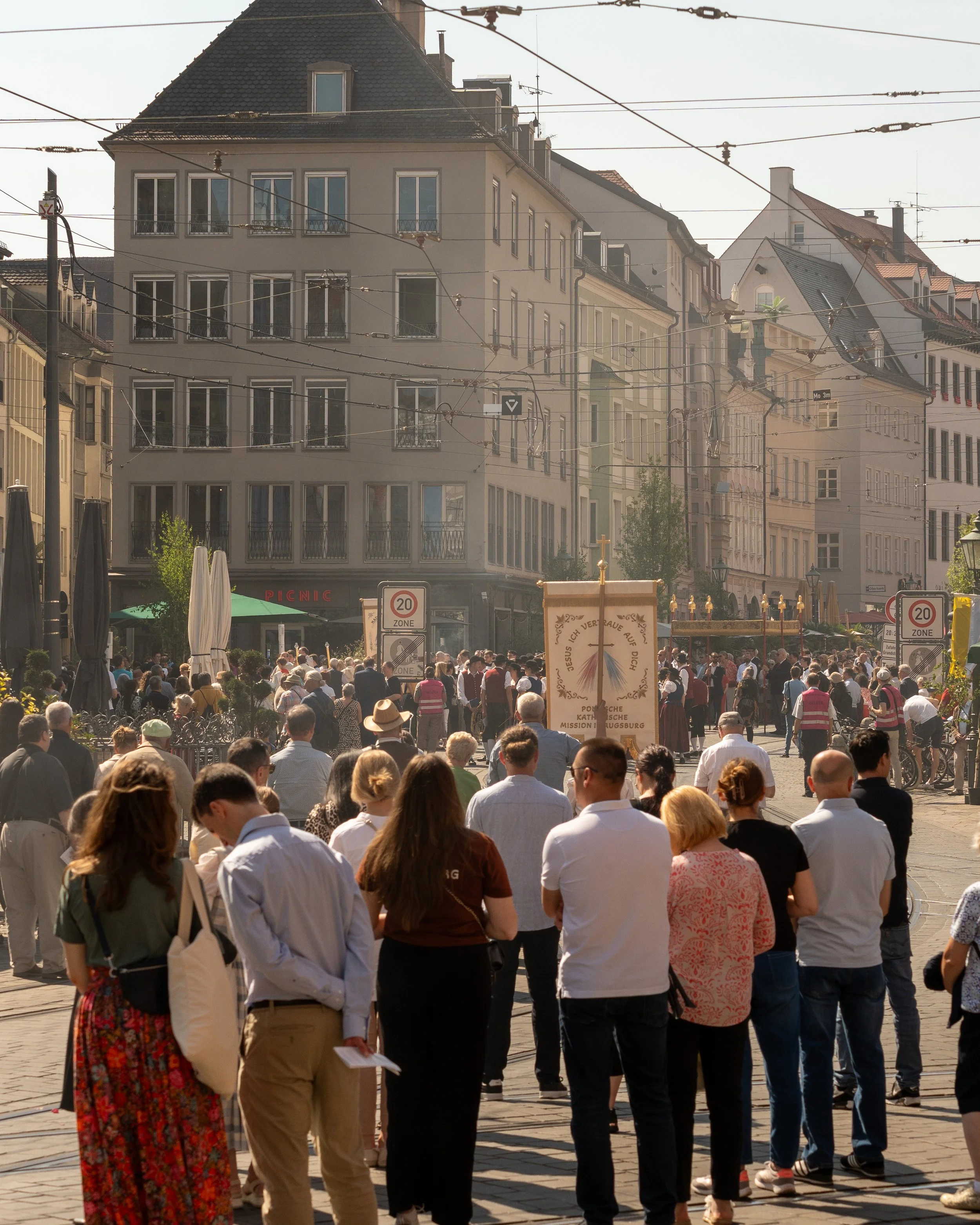 Crowd of people gathered on a city street with tall buildings in the background, some signs indicating a 20 zone, streetcars overhead, and a procession banner with religious text.
