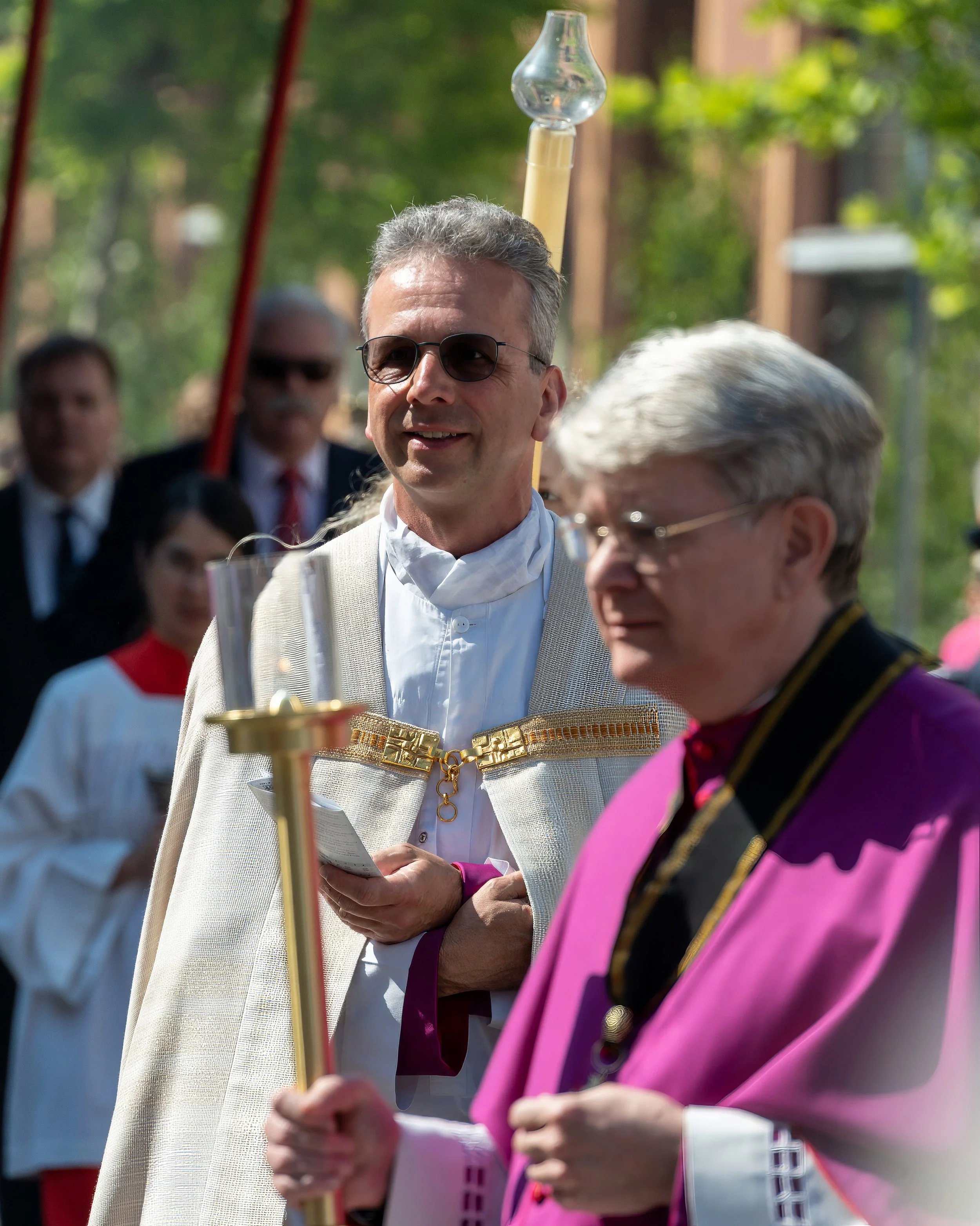 Two men, one in white robes with gold accents and a cape, and the other in vibrant purple and black clergy attire, stand outdoors during a formal event or procession. The man in white is smiling and wearing sunglasses, while the other man appears foc