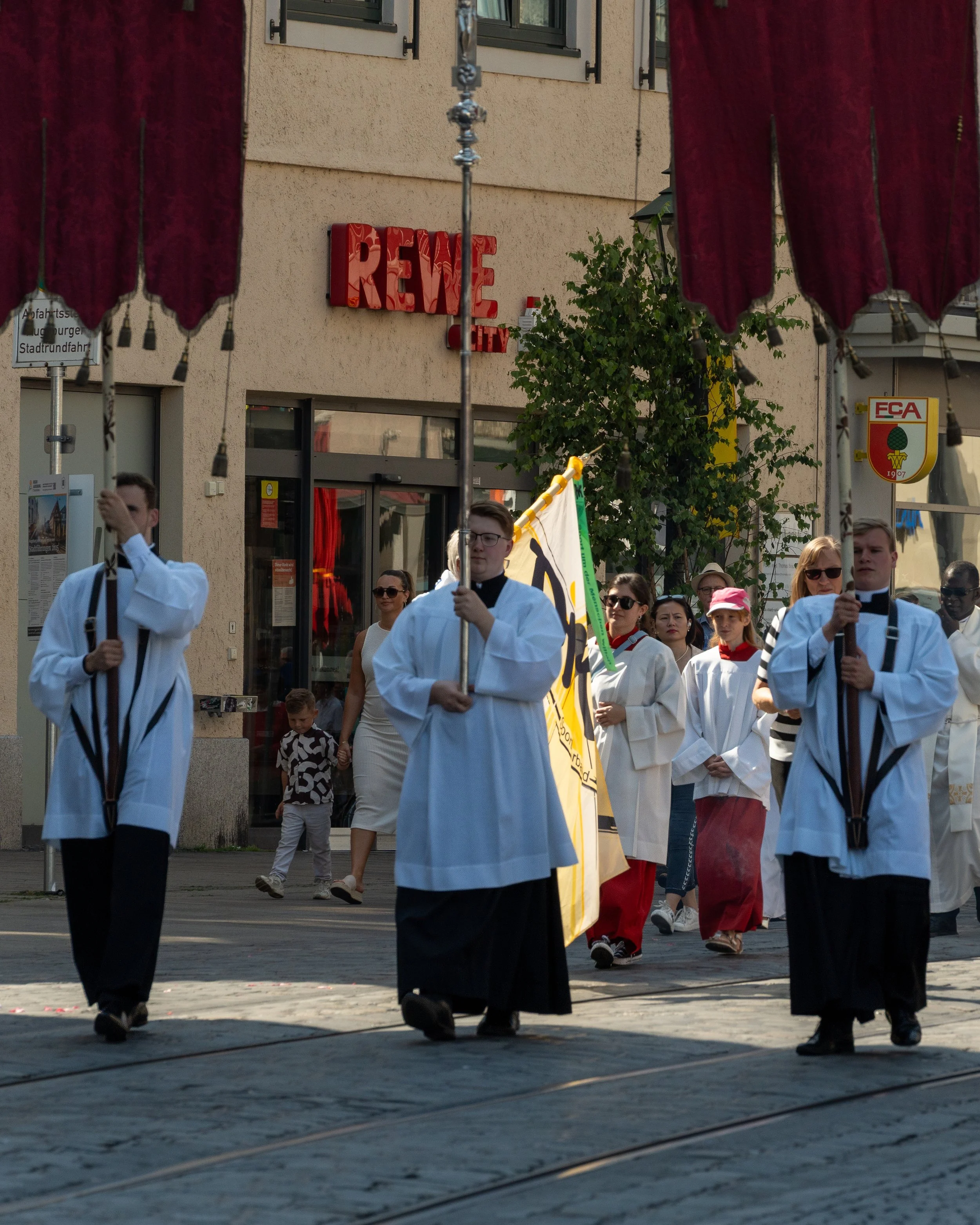 People participating in a procession, some dressed in religious robes, carrying flags, walking on a street with tram tracks, with a red "REWE" store sign in the background.
