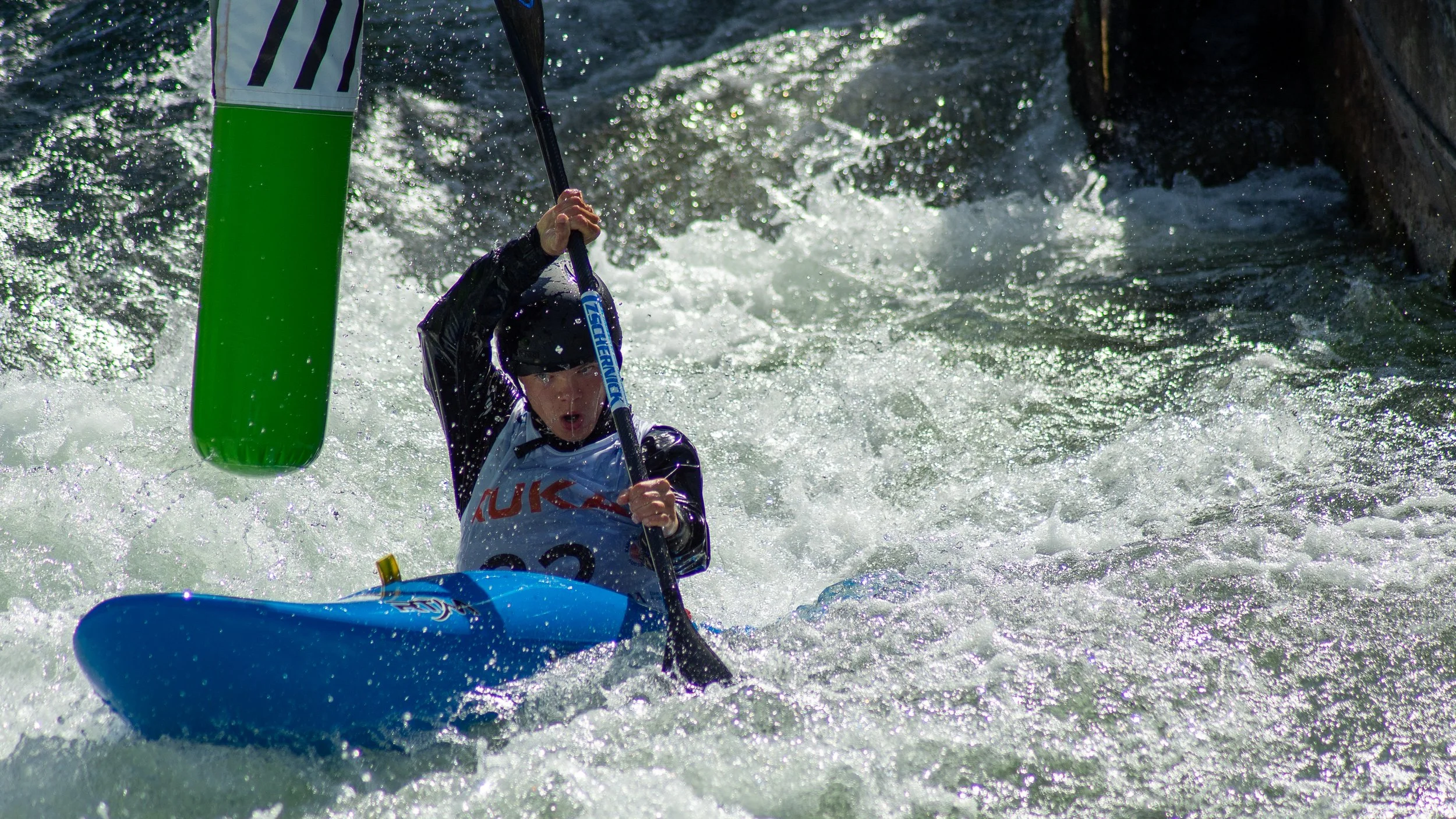 A kayaker wearing a black helmet and black jacket paddles through rough white water rapids in a blue kayak, with an intense expression.