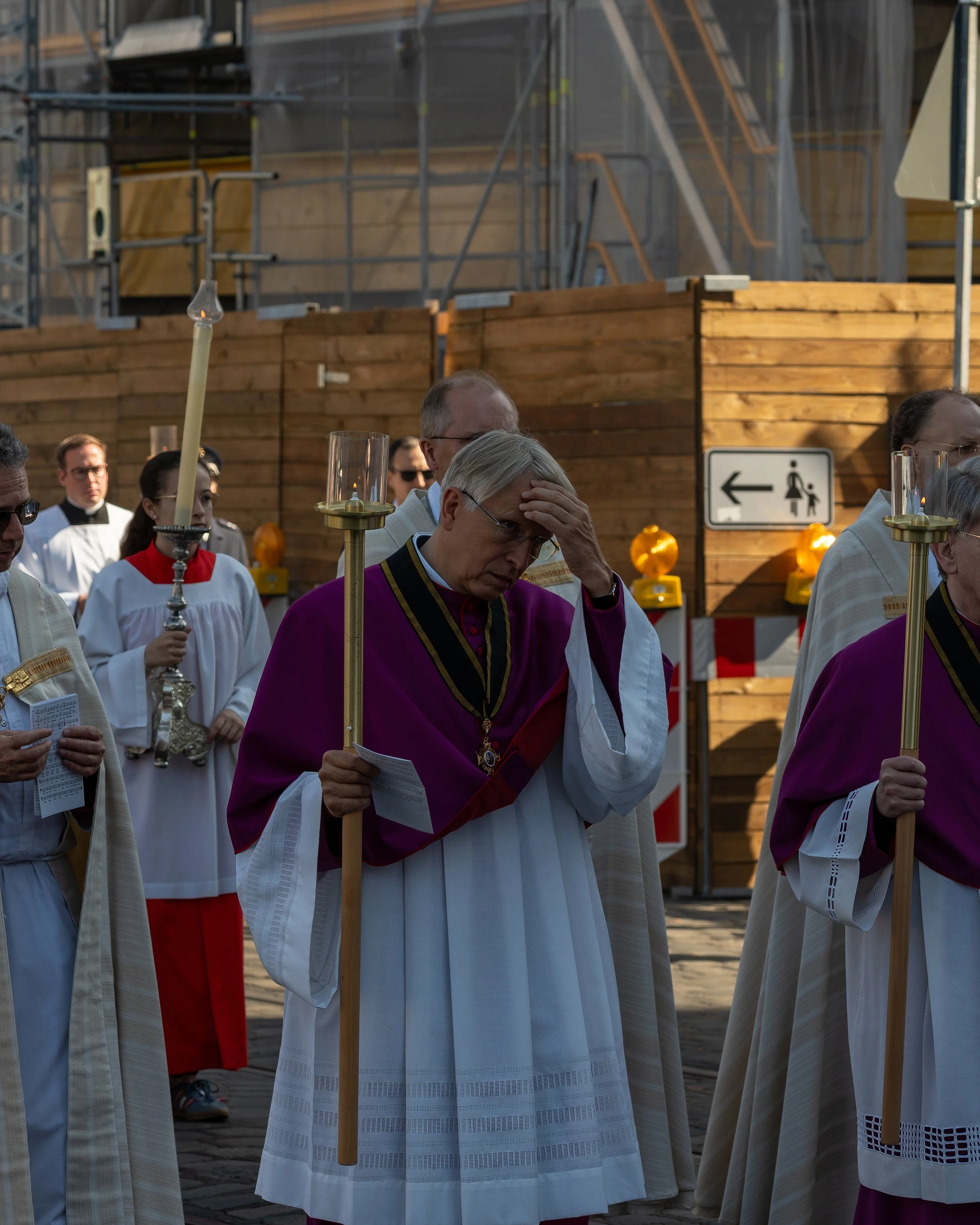 A group of religious figures, including a priest dressed in purple and white robes, holding candles and prayer books, participating in a religious procession or ceremony outdoors.