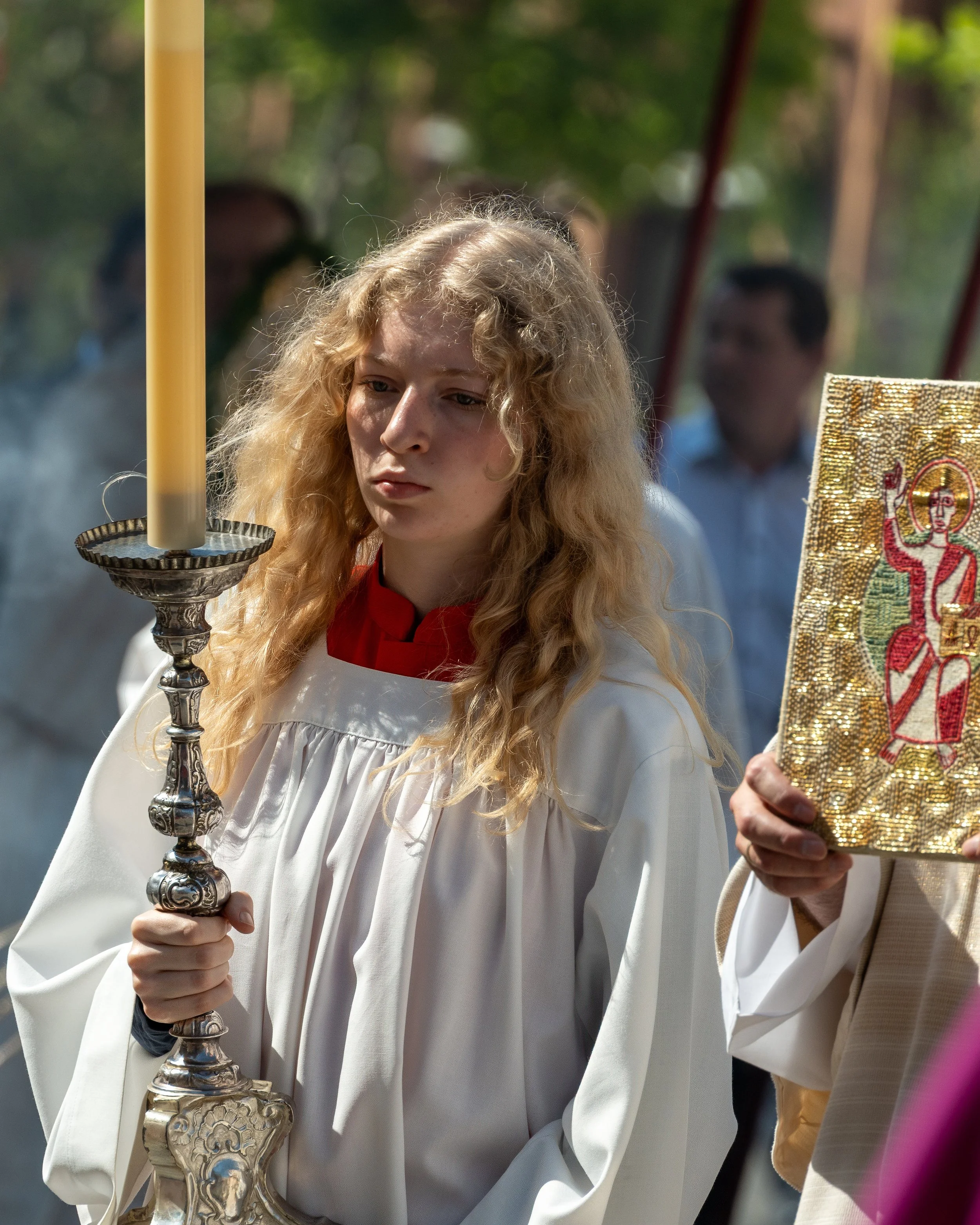 A young woman with long, curly blonde hair wearing a white religious robe, holding a tall candlestick with a lit candle during a religious ceremony outdoors.